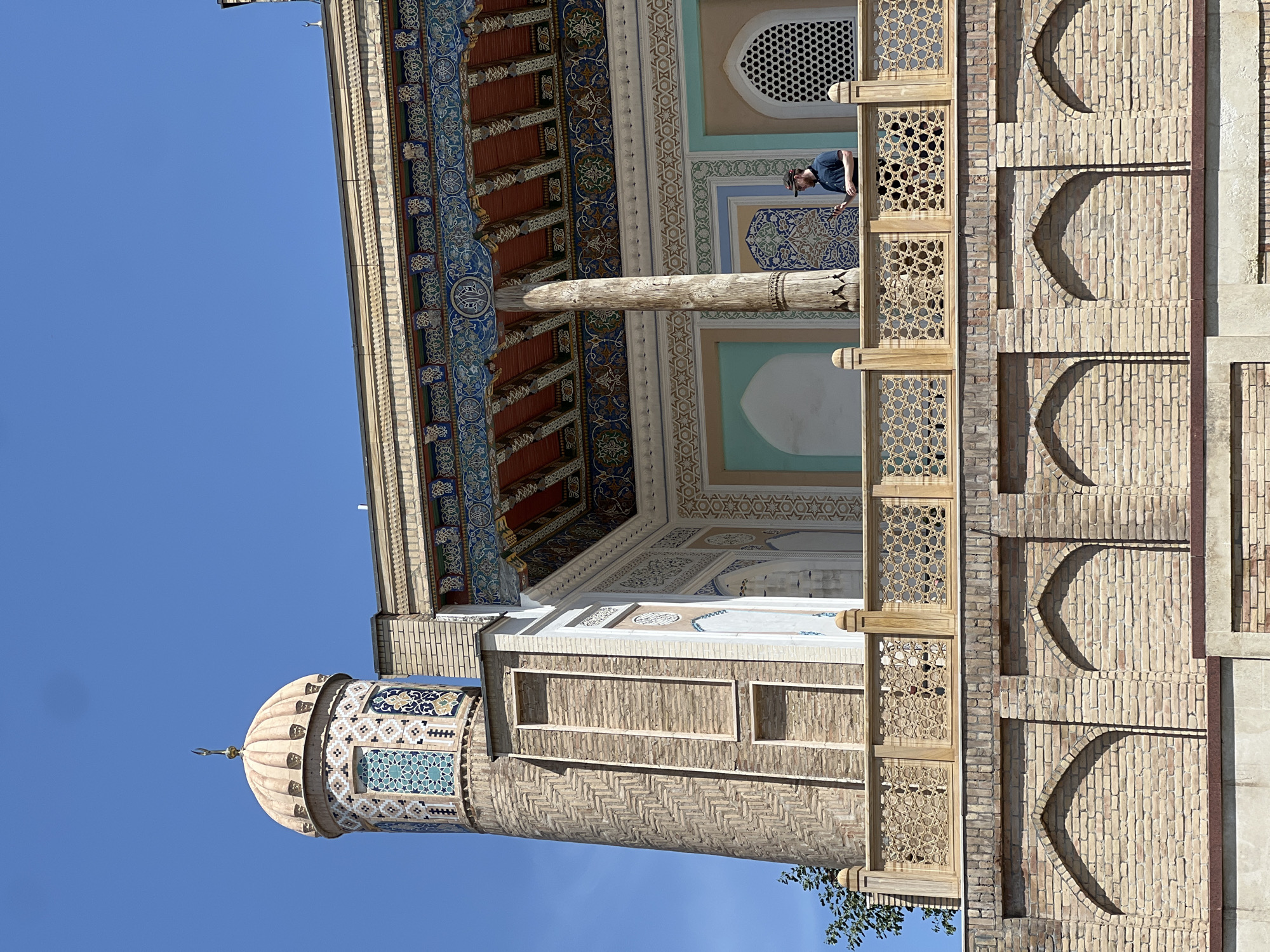 A man stands on a balcony of a building in Samarkand City, Uzbekistan, with a minaret and dome visible in the background. The architecture features intricate tilework and decorative brickwork. The man is wearing a dark shirt and a baseball cap and appears to be looking at something off-camera. The sky is clear and blue.