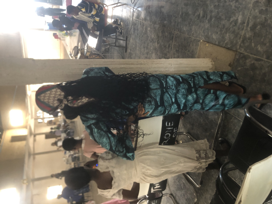 An indoor gathering is depicted within a large, light-walled hall in Samaru, Nigeria. In the immediate foreground, a woman faces away from the camera, dressed in a form-fitting teal gown with a dark, leaf-like pattern. She has long black braids and wears a patterned headscarf displaying red, green, and cream colors, possibly with a repeating "DIOR" logo. Her hands are on her hips as she leans slightly against a light-colored concrete pillar. To her left, a second woman, also facing away, wears a light-colored, possibly off-white, lace dress. A white sign on a stand between them partially displays the text "RONE OF". Several modern chairs with dark seats and chrome frames are in the foreground. In the midground on the right, two young men are seated; one plays a black electronic keyboard, while the other, wearing a striped yellow and blue shirt, observes. A partial drum set is visible behind the keyboard. Numerous blurred figures are present throughout the brightly lit background, some standing and some seated, indicating a larger congregation. The environment features light-colored walls, a tiled grey floor, and strong natural light entering from openings or windows in the background.