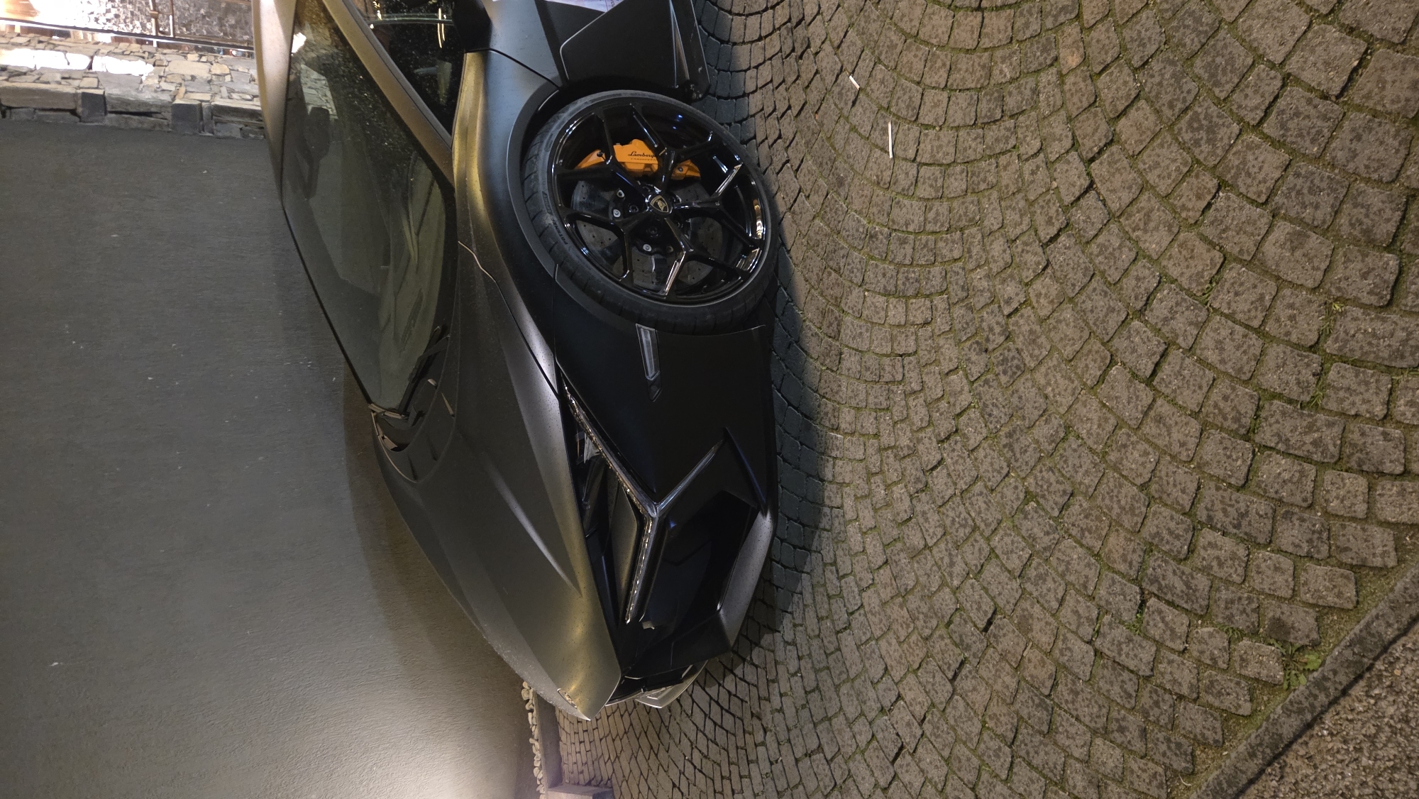 Here is a description of the image:
A portion of a matte gray Lamborghini sports car occupies the frame's left side, positioned on a pavement composed of gray cobblestones. The car features a black wheel with orange brake calipers and a distinctive front headlight design with a Y-shaped LED accent. The pavement appears slightly damp, with hints of greenery between the cobblestones, suggesting a well-maintained urban environment. The backdrop includes a dark, flat surface and a stone wall detail that suggests an urban street setting. Overall, the composition and lighting create a high-contrast image highlighting the car's design.