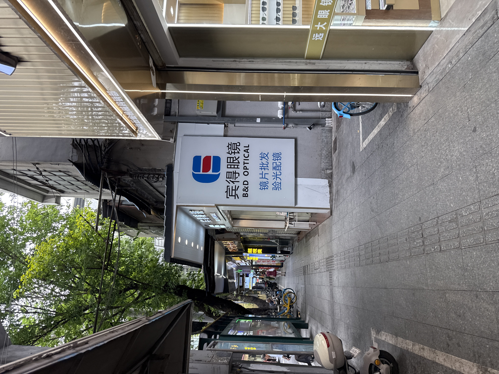 The image shows a narrow pedestrian street in Yuexiu District, China, lined with shops and buildings. The street is paved with gray stone tiles, and there are tactile paving indicators along the center. On the right side of the street is a shop with a golden facade and large glass windows displaying eyeglasses. A sign above the window reads "B&D OPTICAL" in English and Chinese characters. Further down the street, there are more shops with illuminated signs, including one with the characters "远大眼镜" which translates to "Yuanda Eyeglasses". On the left side of the street, there are awnings extending from buildings, a bus stop sign, and a few parked motorcycles and bicycles. A tree with lush green foliage grows between the buildings, casting shadows on the walkway. A woman in a red dress is walking away from the camera in the distance. The sky is overcast.