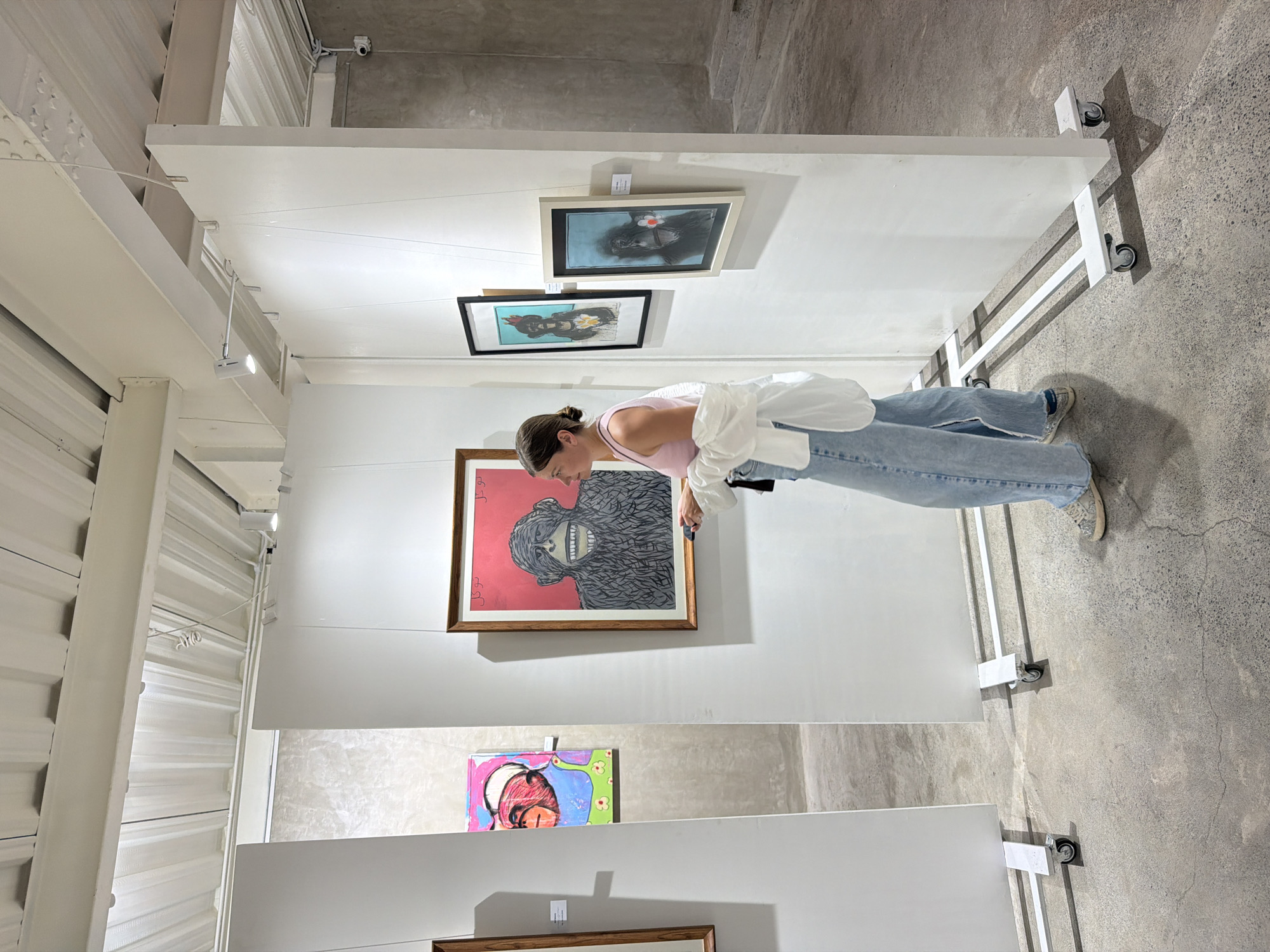The image depicts a woman in a pink tank top and blue jeans looking at artwork displayed in a gallery. The gallery appears modern with white walls and concrete floors. There are several framed pieces of art on display, including a large painting of a monkey-like creature with a red background and a smaller colorful abstract piece. The woman is leaning forward, appearing engaged with the artwork. The lighting suggests an indoor setting, possibly daytime given the brightness. The overall mood seems relaxed and contemplative, typical of an art gallery visit. The location is Kuta, Indonesia.