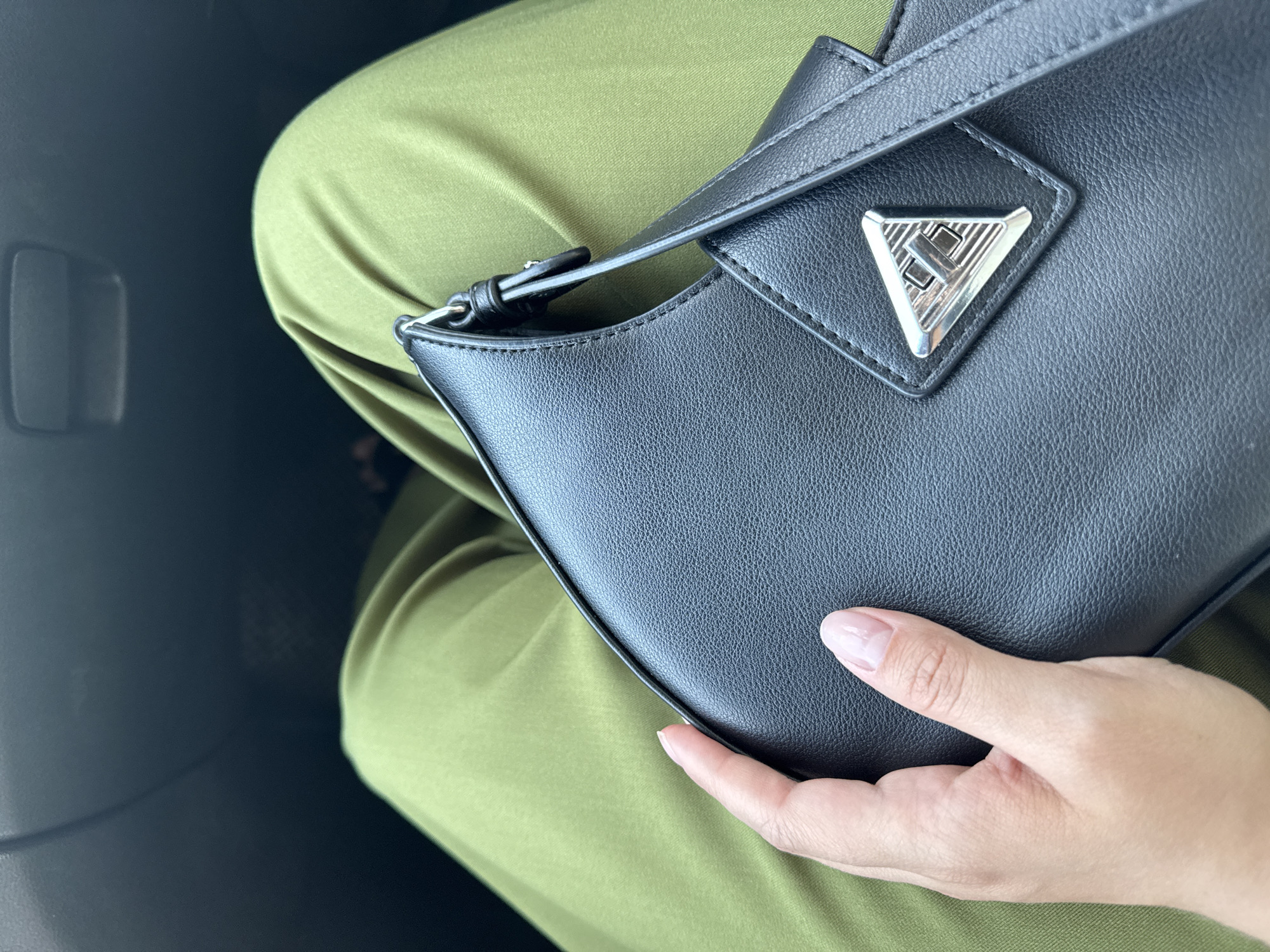 A person's hand is holding a black handbag with a silver triangular logo. The person is wearing olive green pants. The background appears to be the interior of a car. The scene is not in Chaikovsky, Russia.
