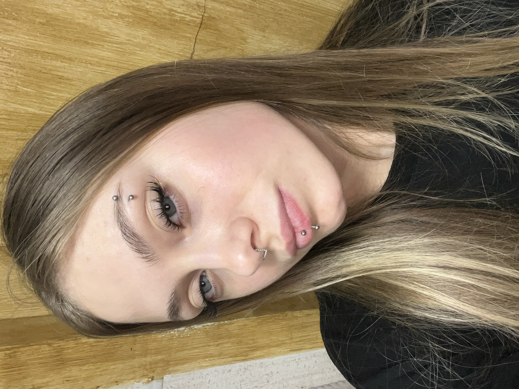 A medium shot features a young woman with fair skin, light brown hair with blonde highlights, and blue eyes. She has multiple facial piercings, including a horizontal eyebrow piercing on her right eyebrow, a nose piercing, and a lip piercing on the lower lip. She is wearing a black shirt. The background consists of a wooden surface. There is no indication of the city of Фоки, Russia in the image.