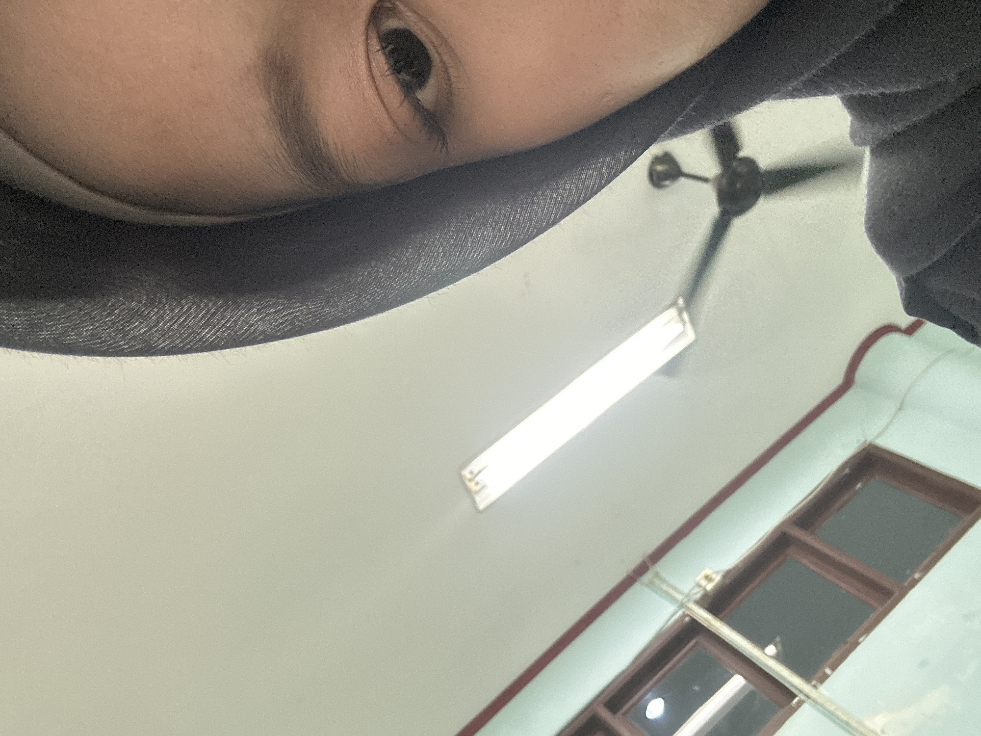 The image is a close-up, angled shot of a person's face, specifically their right eye and part of their forehead, partially obscured by a dark grey hijab. The background is an indoor environment, appearing to be a room with pale green walls. A fluorescent light fixture is visible on the ceiling, along with a dark ceiling fan that is slightly out of focus. Two windows with dark brown frames are also visible on the wall, with reflections of light on their surfaces. The image is taken from a low angle, looking upwards. The location is identified as Reuleut Barat, Indonesia.
