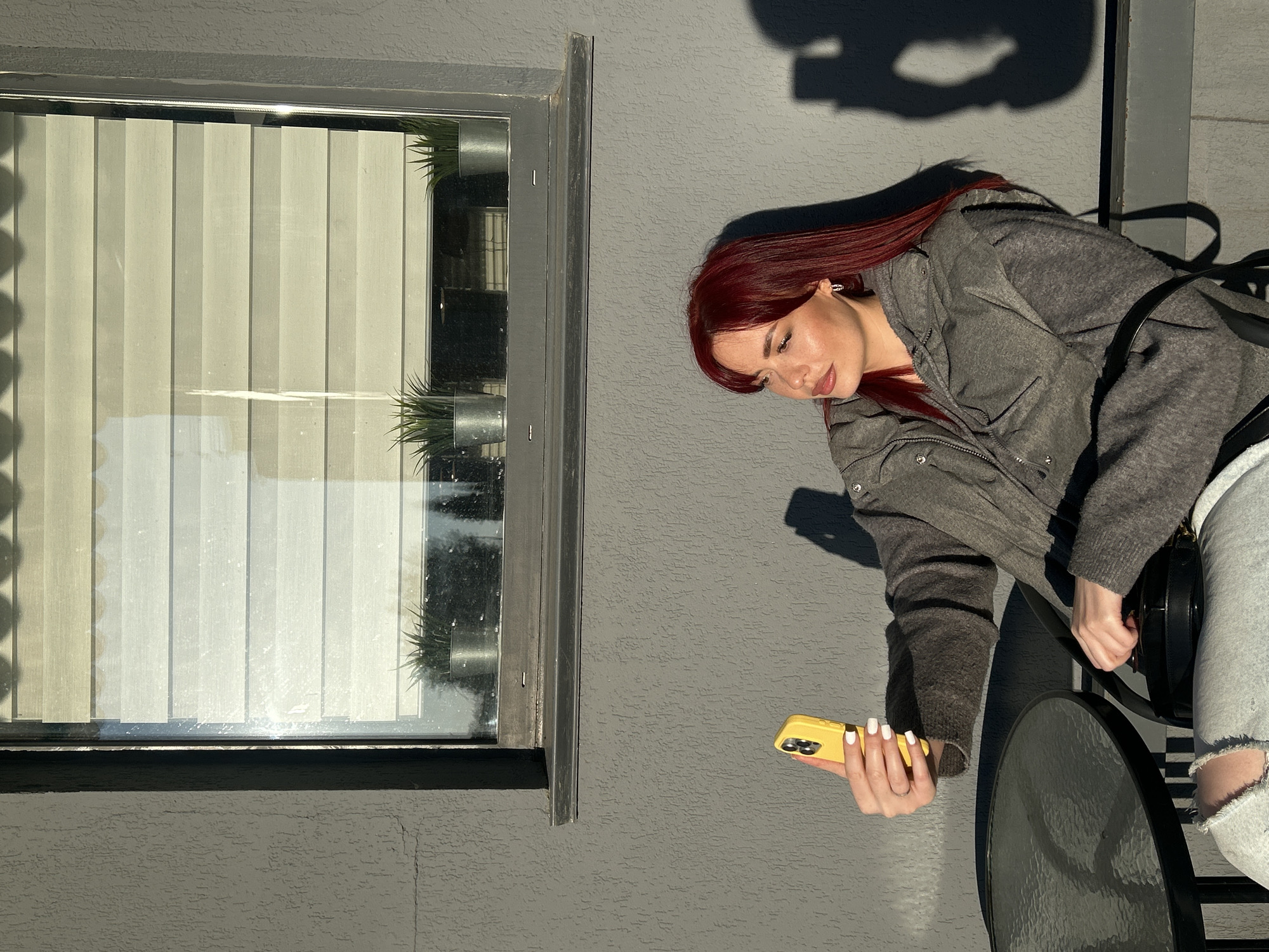 The image depicts a young woman outdoors on a balcony or patio in Tolyatti, Russia, focused on her smartphone. The lighting suggests either early morning or late afternoon, with strong, elongated shadows cast by direct sunlight on a textured grey wall.
The woman has long, straight, vibrant red hair and is shown in profile, looking down at her yellow-cased smartphone, held in her right hand with white-painted fingernails. She is wearing a grey jacket or cardigan and light-colored jeans, which are visibly ripped at the knee. A black shoulder bag is slung over her. She is leaning over a round table with a glass top and a dark metal frame.
To her left, a window with a dark frame is set into the grey wall. Through the window, vertical blinds are visible, along with two small potted plants (possibly artificial) placed on the interior windowsill. The scene conveys a moment of calm, with the woman engrossed in her device amidst the bright, natural light. No discernible text is visible on the phone screen or elsewhere in the image.