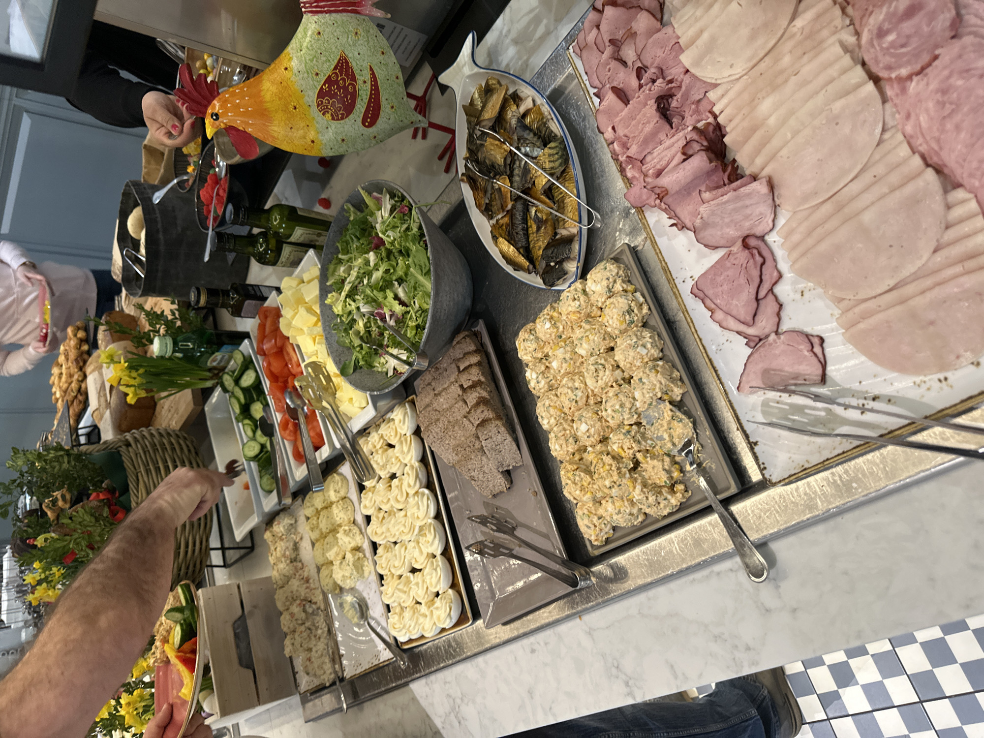 The image shows a buffet table with various food items, likely in Wrocław, Poland.
From left to right, the items include:
- Assorted bread and pastries in a wicker basket.
- Vegetables like cucumbers and tomatoes.
- A platter of deviled eggs.
- Sliced brown bread.
- A tray of salad with mayonnaise and eggs.
- A fish-shaped dish filled with smoked fish.
- Several types of sliced deli meat.
There are people at the buffet, one appearing to be serving themselves vegetables with a fork. There is a decorative ceramic chicken near the center. The table surface is made of marble, and the floor has a blue and white checkered pattern.
