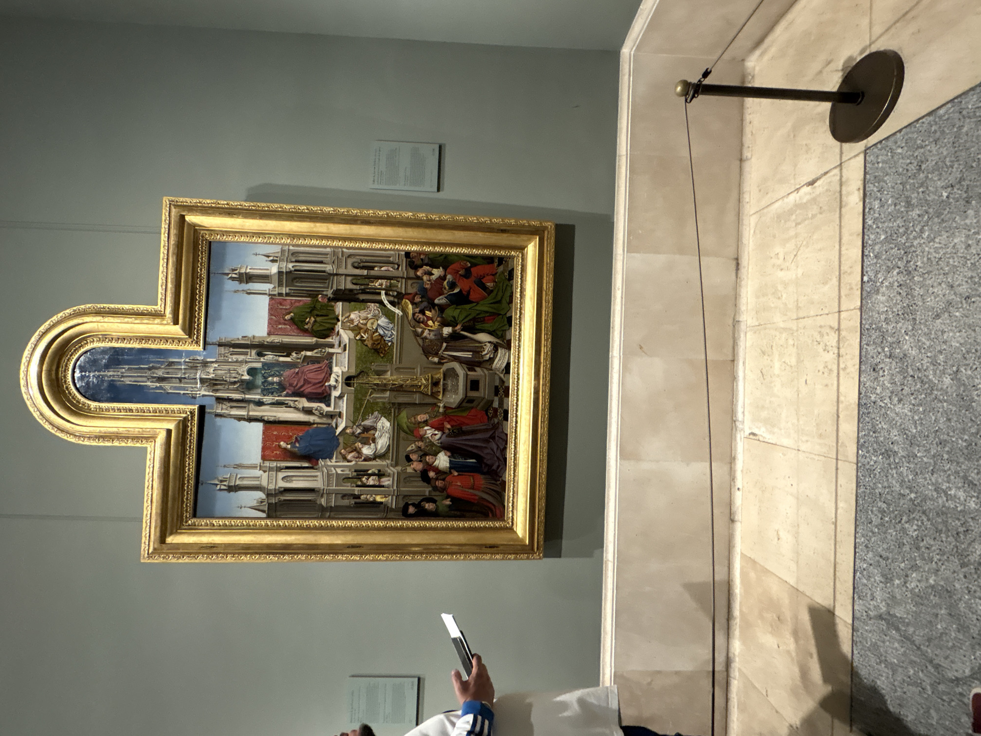 The image shows a painting in the Museo Nacional del Prado, located in Madrid, Spain. The painting, framed in gold, depicts a historical or religious scene with multiple figures in elaborate clothing and architectural settings. A person holding a smartphone is partially visible at the bottom of the frame, seemingly taking a picture of the painting. To the right of the painting is a section of the museum wall with marble or stone paneling and a security barrier.