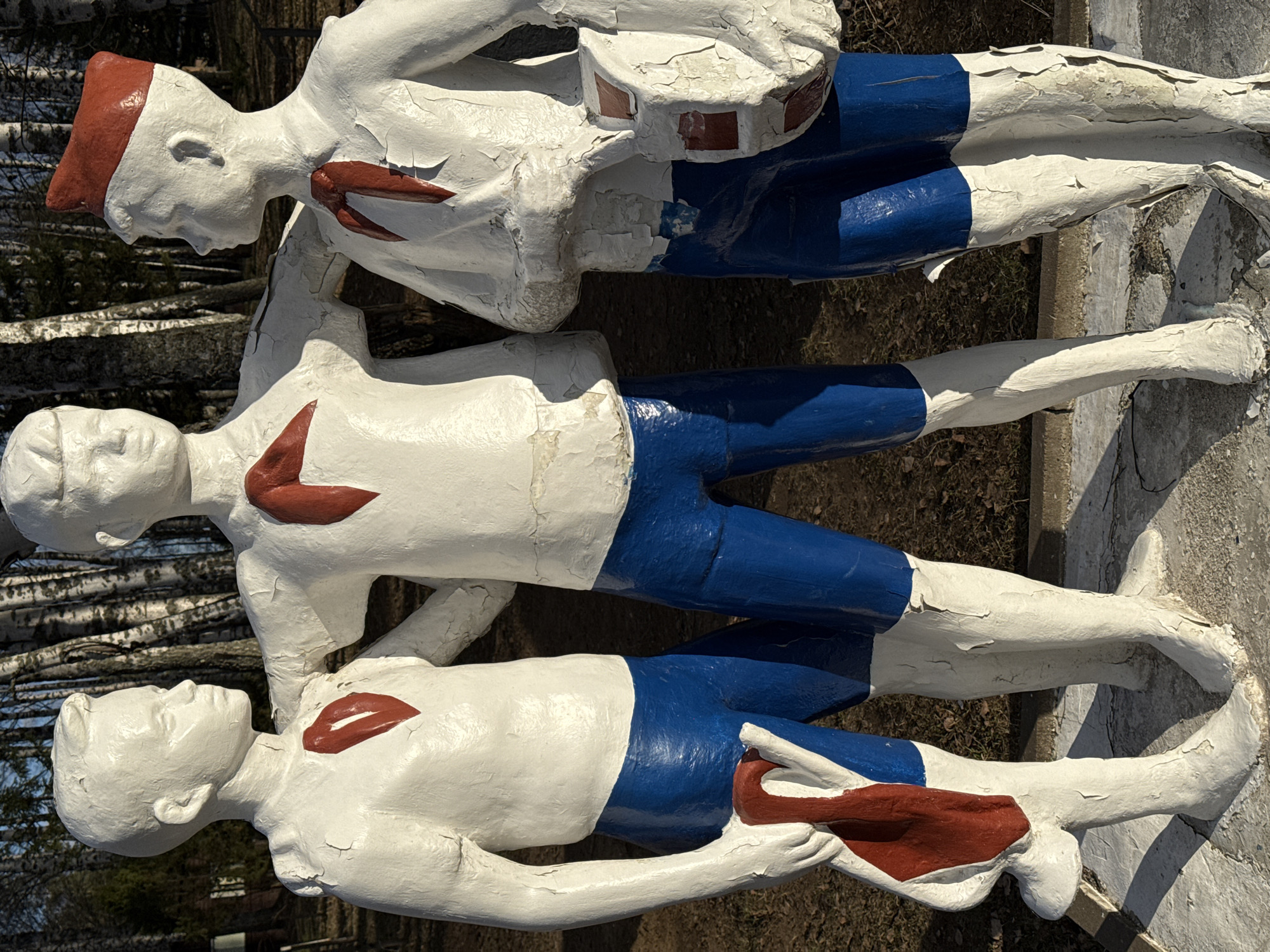 The image shows a sculpture of three male figures in Galevo, Russia. The figures are arranged vertically, one on top of the other. They are painted white, with red markings on their chests and red headwear on the top figure, and blue shorts. The paint is cracked and peeling. The background includes birch trees and ground cover. The sculpture sits on a concrete base.