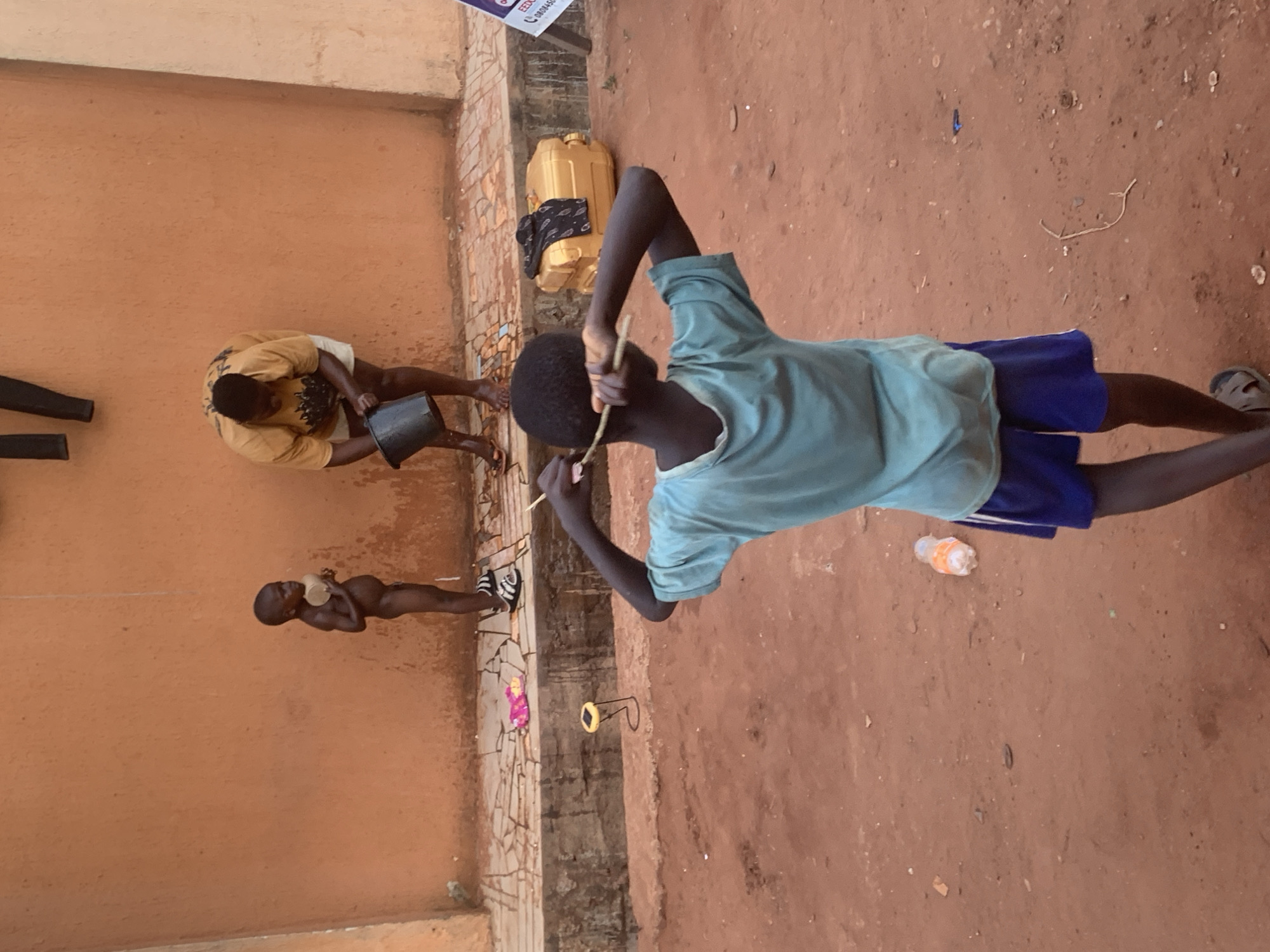 The image shows a scene in Ukwu-Abwa, Nigeria. Three individuals are visible. The central figure, a child in a light blue shirt and dark blue shorts, stands on reddish-brown soil. He holds a thin stick and appears to be engaging in some activity. Beside him, there is a transparent bottle with an orange cap.
To the left, near an orange-colored wall, is another child, who has a cup. Further behind, there is another person is pouring something from a bucket. Behind the central figure, a yellow water container is visible against the wall. The ground is dirt and there is an old wall.