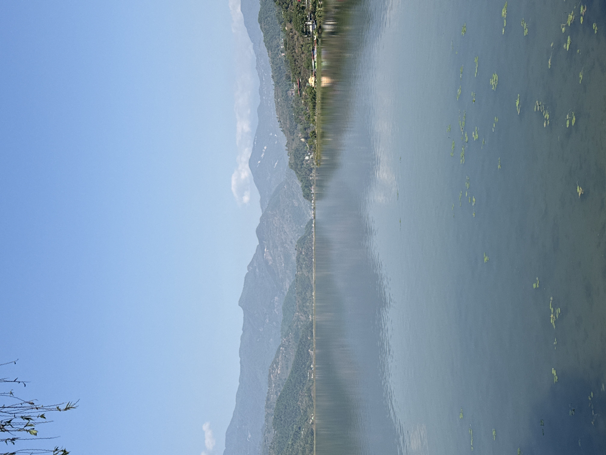 The image depicts a serene landscape in Pokhara, Nepal, likely near Phewa Lake. Mountains covered in green vegetation are visible in the background, reflected in the calm water of the lake, which also has patches of aquatic plants floating on its surface. The sky is clear and blue.