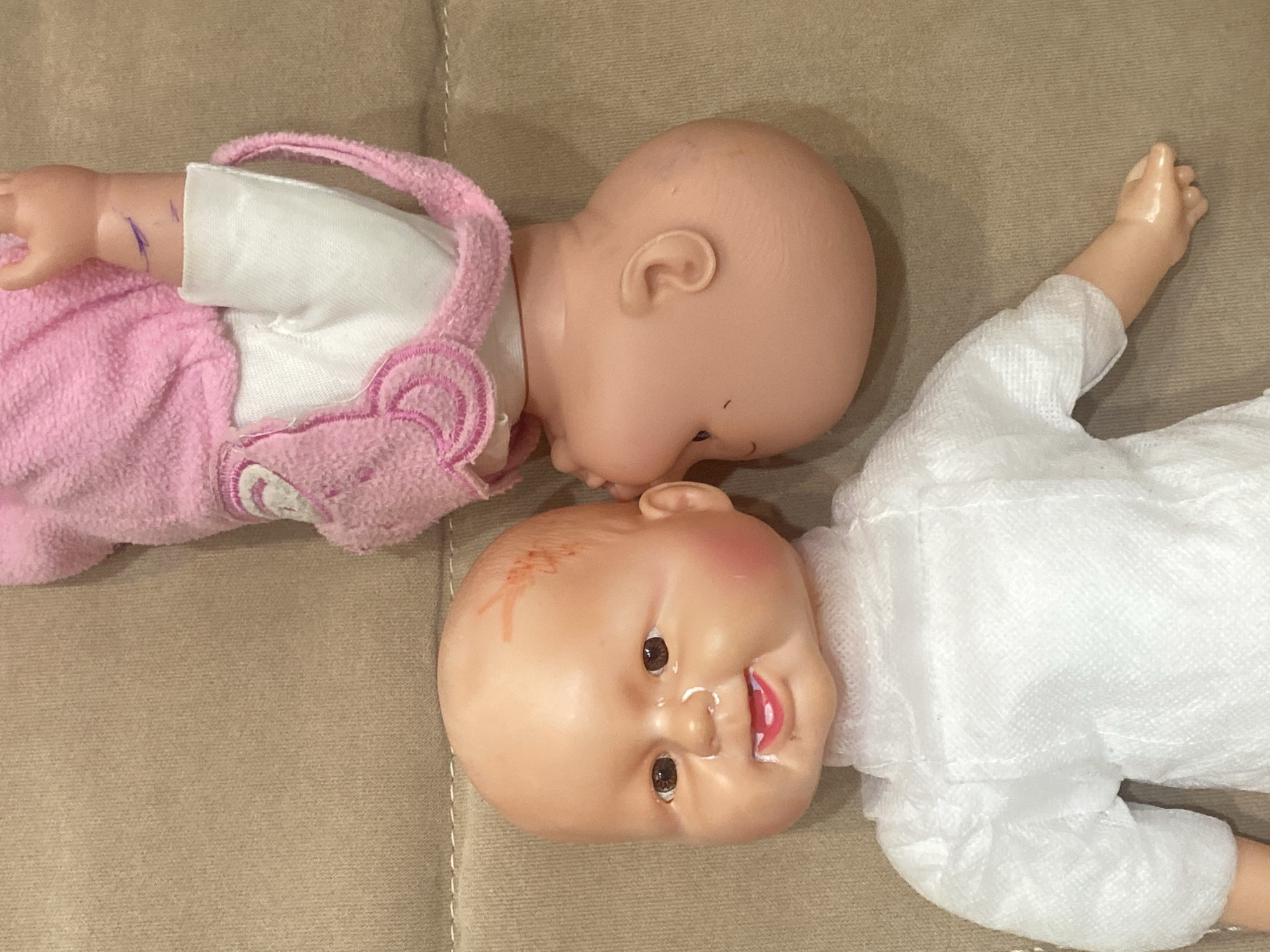 Two baby dolls are positioned on a beige, textured surface, which appears to be a sofa or carpet. One doll, in the foreground and oriented upright, has a bald, light-skinned head with brown eyes, a slightly open mouth showing teeth and tongue, and rosy cheeks. It is wearing a white, textured garment with puffed sleeves. There are some faint red marks on its forehead. The second doll, in the background and upside down, is wearing a pink hooded garment with white trim. Its head is turned away from the viewer, with only its ear and the back of its head visible. Purple marks, possibly from marker, are visible on its arm. The overall scene is brightly lit, suggesting an indoor setting. There is no information available to determine the location of this scene in the city of Dolgoprudny, Russia.