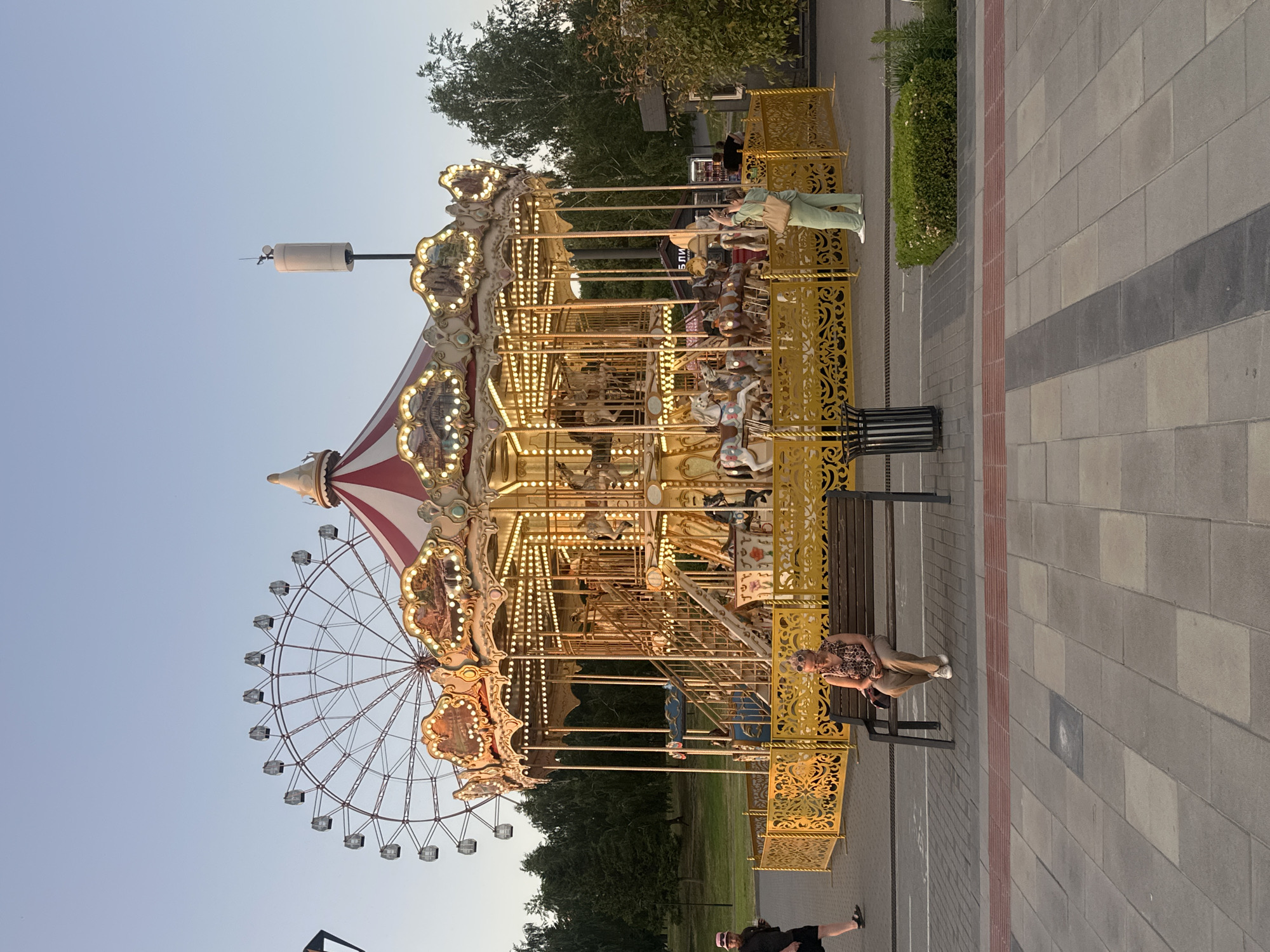 The media file captures an outdoor amusement park scene with a carousel as the central subject. The carousel is brightly lit with numerous lights, suggesting it is either late afternoon or early evening. It is a classic design with ornate golden railings and decorative panels, featuring various carousel animals and carriages for riders. A large Ferris wheel is partially visible behind the carousel, its structure hinting at a fairground or amusement park setting.
Several people are present in the scene. A woman in a light green outfit is standing near the carousel entrance, appearing to interact with someone or observe the ride. On a bench next to the carousel, a woman wearing a leopard-print top and light-colored pants is seated, looking towards the carousel with a relaxed posture. Another person is partially visible in the foreground, suggesting ongoing activity in the park. The ground is paved with rectangular gray and red bricks, indicating a public outdoor space. Lush green trees and bushes surround the amusement park structures, adding to the outdoor setting. The sky is a clear, pale blue, typical of a calm, possibly twilight, atmosphere. The overall mood is one of leisure and entertainment. No discernible text is visible in the image. The location context provided is Novosibirsk, Russia.