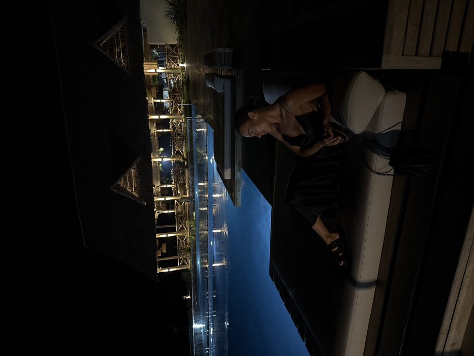 A woman sits on a cushioned outdoor lounge next to an infinity pool at night. She wears a black dress and sandals. The pool reflects the lights from a nearby open-air building with a thatched roof. The scene is dark with illuminated areas highlighting the structures and reflection.