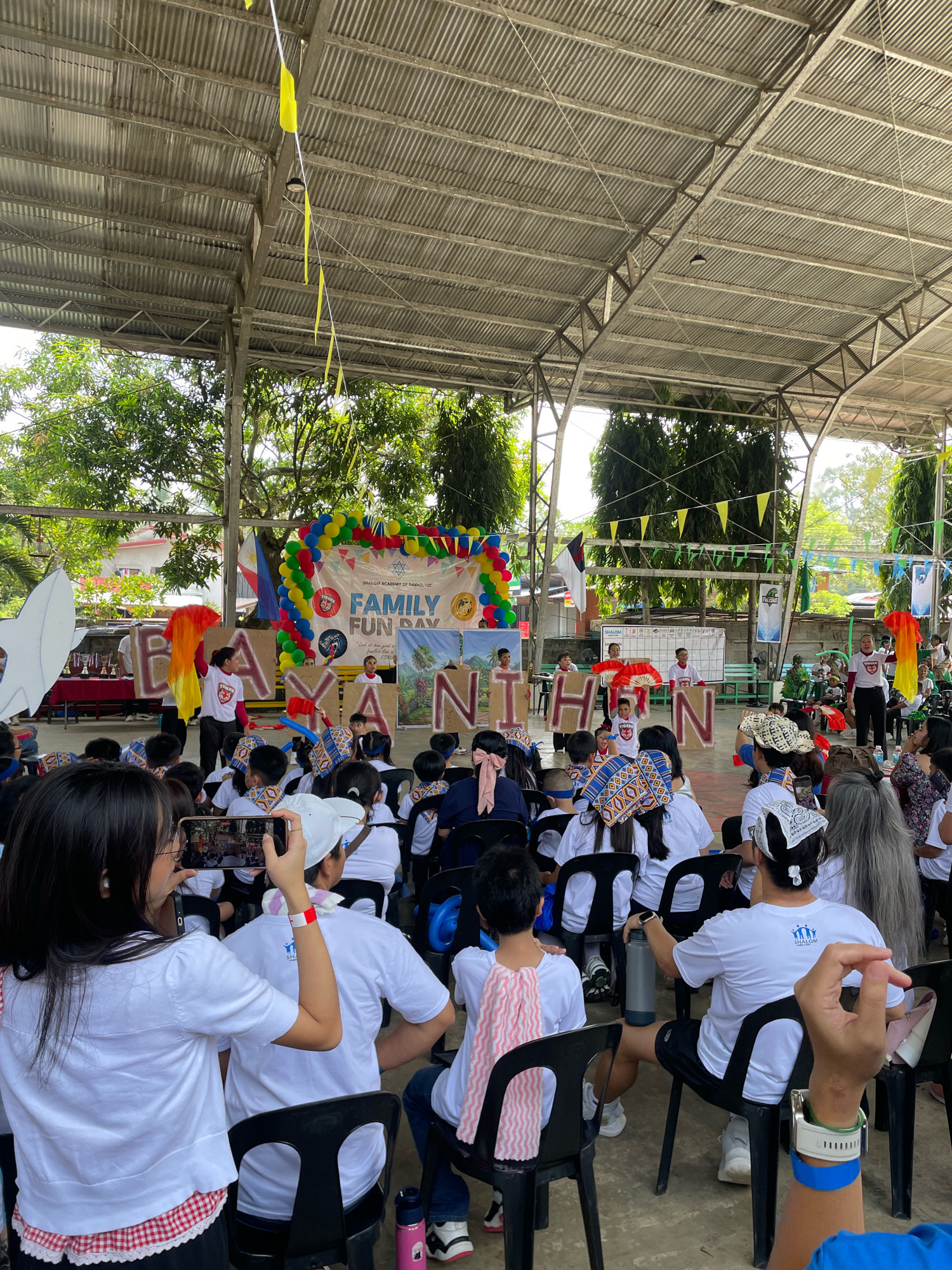 This is a medium shot, taken from behind and to the side of the audience, of a "FAMILY FUN DAY" event held outdoors under a corrugated metal roof supported by metal poles, likely in Davao City, Philippines. The event appears to be a school-organized activity, as indicated by the banner that reads "SHALOM ACADEMY OF DAVAO, INC. FAMILY FUN DAY" and "BAYANIHAN."
In the foreground, several audience members are seated on black plastic chairs, facing a raised stage area. Many of them are wearing white t-shirts, some with logos. One person on the left is holding up a smartphone, recording the event. Another person's hand, wearing a blue wristband and a watch, is visible in the bottom right corner.
On the stage, a group of children and adults are participating in a performance. Some children are holding large, brown cardboard letters that spell out "BAYANIHAN." Two individuals are holding red fans and performing a dance. Another person is holding a large, orange and red fan-like object. A banner with balloons and a Philippine flag is visible behind the performers.
The environment includes trees with green foliage visible in the background, suggesting an outdoor setting. The overall atmosphere is festive and celebratory, with decorations such as balloons and streamers. The structure of the roof implies it's an open-air gymnasium or a covered court.
