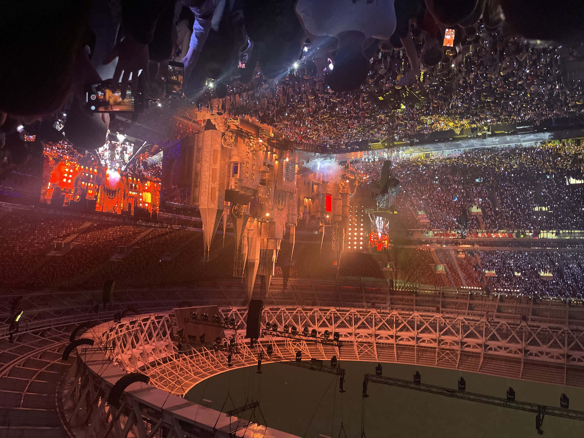 The image depicts a large-scale outdoor concert held at Luzhniki Stadium in Moscow, Russia. The stadium is filled with an enormous crowd of spectators, many of whom are holding up their phones, some with their flashlights on, illuminating the dark space. The stage setup is elaborate, featuring a massive, illuminated castle-like structure with multiple towers and battlements, bathed in warm, orange light. There are also large screens displaying imagery, likely of the performers, and a significant amount of stage lighting and pyrotechnics. The architectural elements of the stadium's roof and seating tiers are visible, adding to the sense of scale and grandeur. The overall atmosphere is that of an energetic and visually impressive live music event.