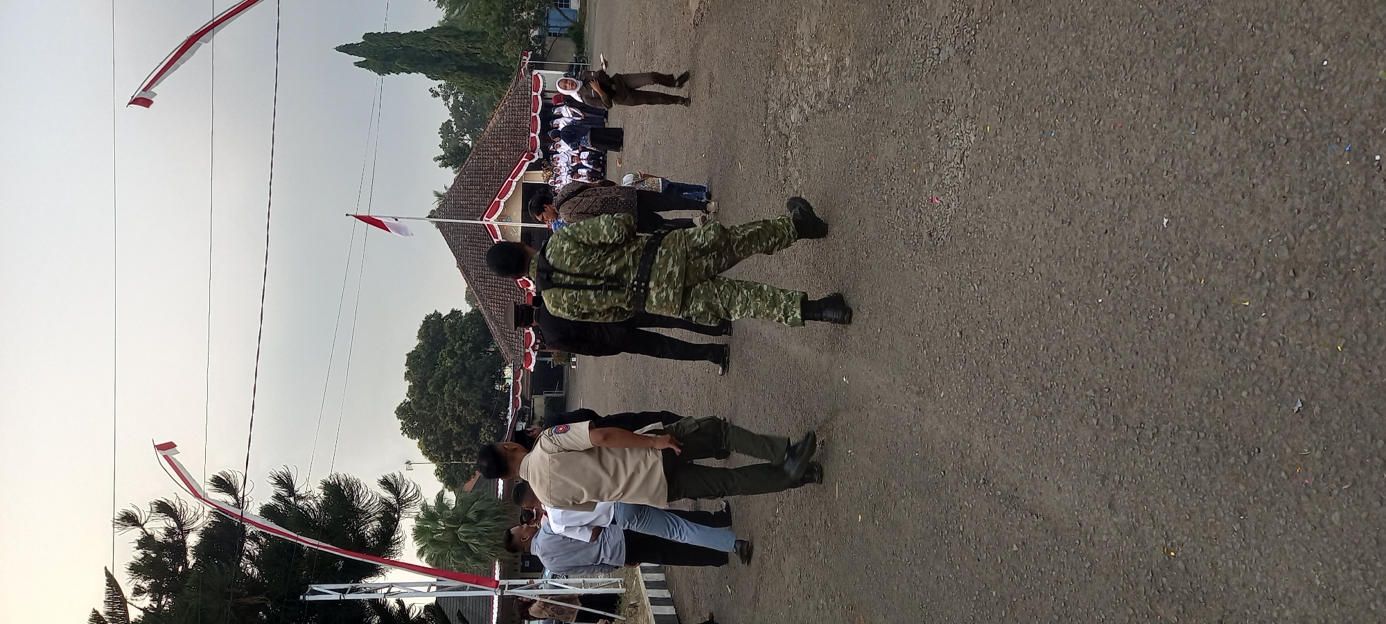 The image depicts an outdoor scene with several people gathered in what appears to be a courtyard or open space. In the foreground, a person in camouflage uniform is walking away from the camera, facing a building with a distinct red and white striped roof and a flag pole flying the Indonesian flag. Several other individuals are seen in groups, some in formal attire and others in what might be school uniforms. The environment is characterized by a paved ground, trees, and utility wires overhead, suggesting an urban or semi-urban setting. The presence of the Indonesian flag indicates a likely national or local event taking place. The building in the background appears to be a communal or administrative structure, possibly a school or government office. The lighting suggests it is daytime, with overcast skies. There is no specific indication of the city of Wanareja, Indonesia in the image itself.