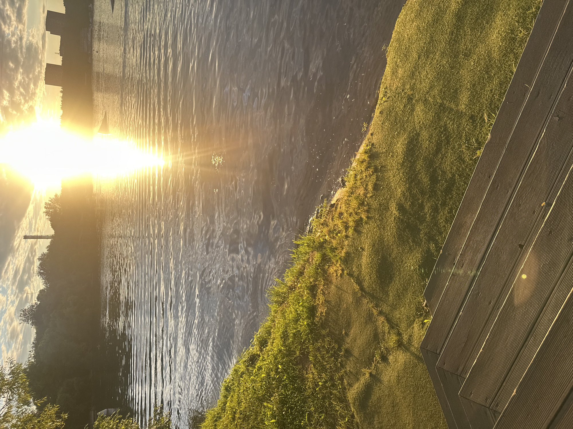 The media file displays a serene waterside scene at sunset in Moscow, Russia. The golden sun, low in the sky, casts a warm, radiant glow across the water, creating shimmering reflections and a soft, diffused light. The water's surface is rippled, suggesting a gentle breeze.
On the right side of the frame, a well-maintained grassy embankment slopes down to the water. Beyond the embankment, a wooden deck or boardwalk with a ribbed texture is visible, indicating a recreational or residential area. The grass is lush and green, with some patches of drier, yellowish grass, suggesting the late summer or early autumn season.
In the distance, across the water, a silhouette of buildings and trees can be observed against the bright sky, hinting at the urban landscape of Moscow. The clouds in the sky are illuminated by the setting sun, adding depth and texture to the scene. A lone sailboat is visible in the water, adding a touch of tranquility and activity to the otherwise still scene.
The overall mood is peaceful and tranquil, evoked by the warm, soft lighting and the natural beauty of the setting. The time of day is clearly sunset, with the sun's low angle and the golden light being prominent features. No people are visible in the media file. There is no visible text.
