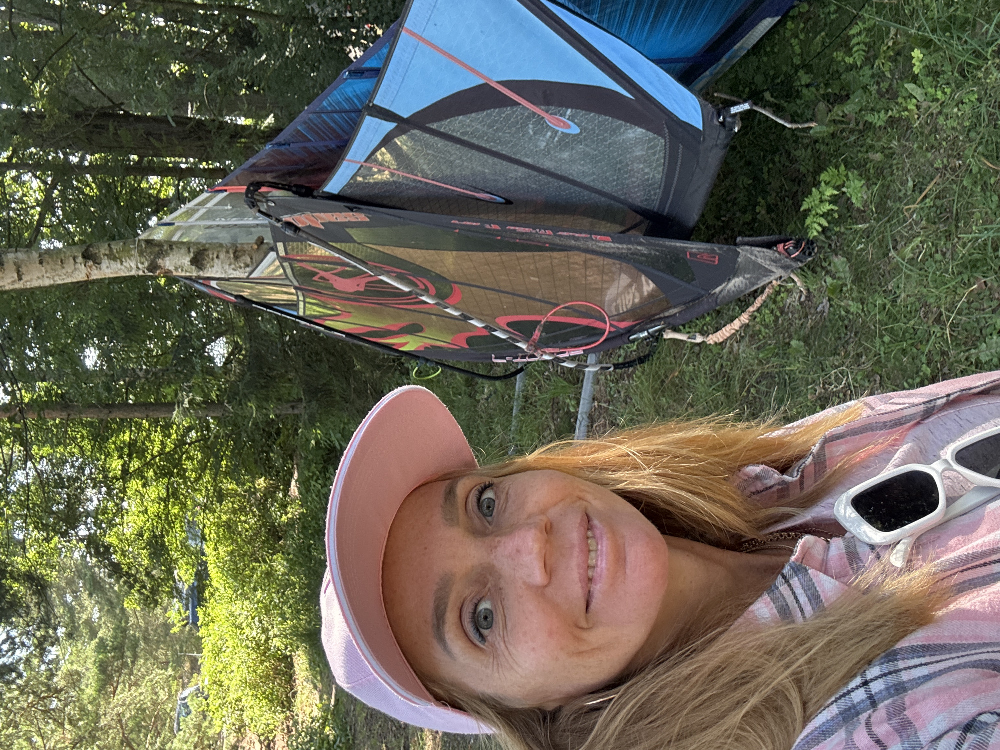 In Zelenaya Roshcha, Russia, a woman is pictured taking a selfie outdoors. She is wearing a pink baseball cap, a pink and white plaid shirt, and a necklace. White sunglasses are tucked into her shirt. She has blonde hair and is smiling at the camera. Behind her, several colorful kiteboarding kites are propped up in a grassy area with trees. The environment appears to be a wooded park or natural area.