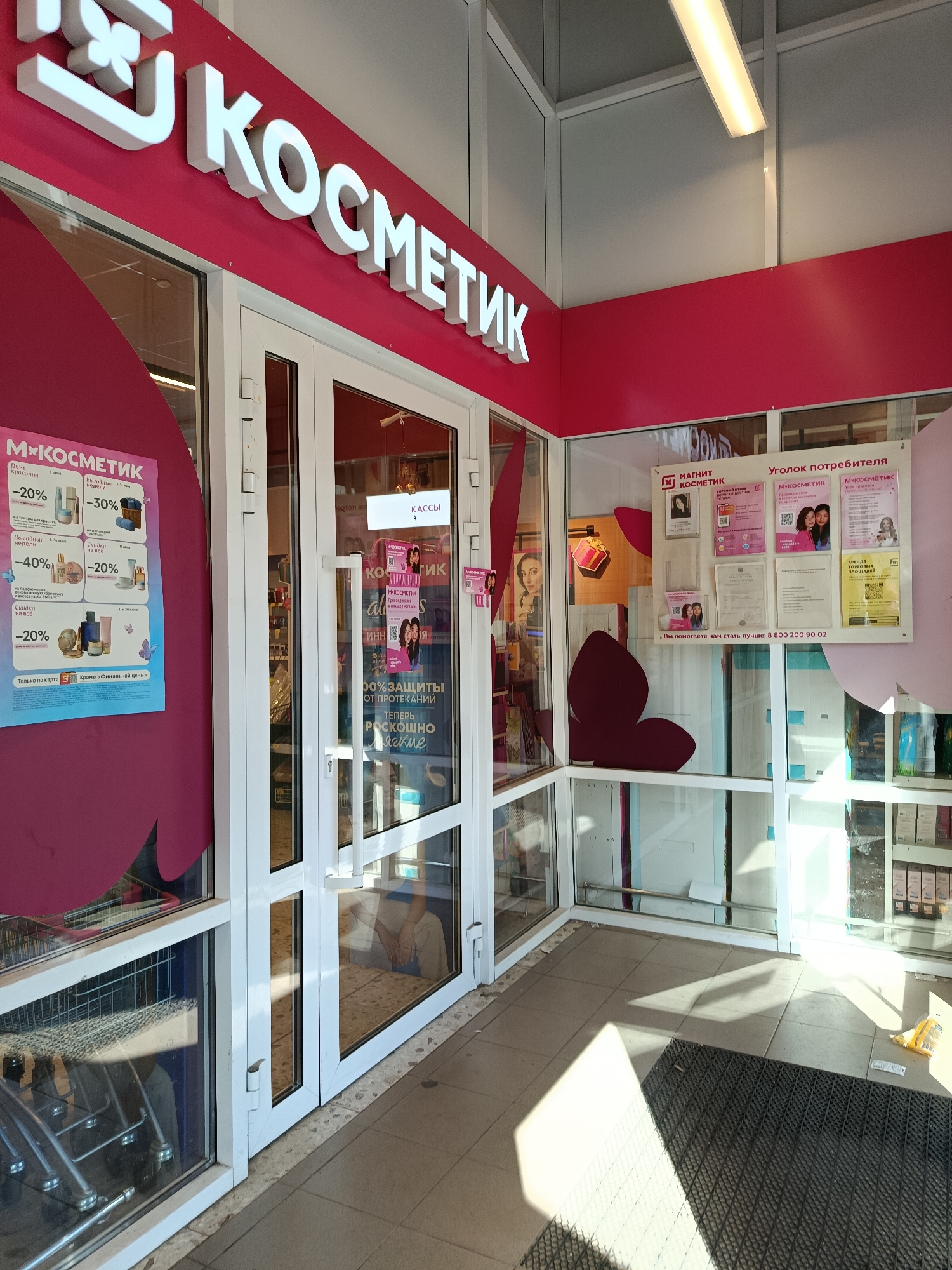 This media file captures the storefront of a "Magnit Kosmetik" store in Orekhovo-Zuyevo, Russia. The store entrance features large glass doors and windows, with bright signage that reads "КОСМЕТИК" in large white letters against a vibrant pink background. Inside, shelves stocked with various cosmetic products are partially visible.
Promotional posters are displayed on the left side of the entrance, advertising discounts of up to 40% on beauty products, with specific offers like "День красоты" and "Выгодные недели." Another poster inside, visible through the glass, also highlights discounts. To the right of the entrance, a "Consumer Corner" is set up with information boards, including details about the store, customer feedback, and contact information, notably a phone number: "8 800 200 90 02."
The scene appears to be during the daytime, as evidenced by the bright lighting and sunlight casting shadows on the tiled floor outside the store. The overall impression is of a commercial establishment ready for customers. The atmosphere seems neutral, focused on the retail aspect of the business. No specific people are prominently featured, though reflections in the glass might hint at passersby or individuals inside. The setting is clearly a street-level retail space in a commercial area.