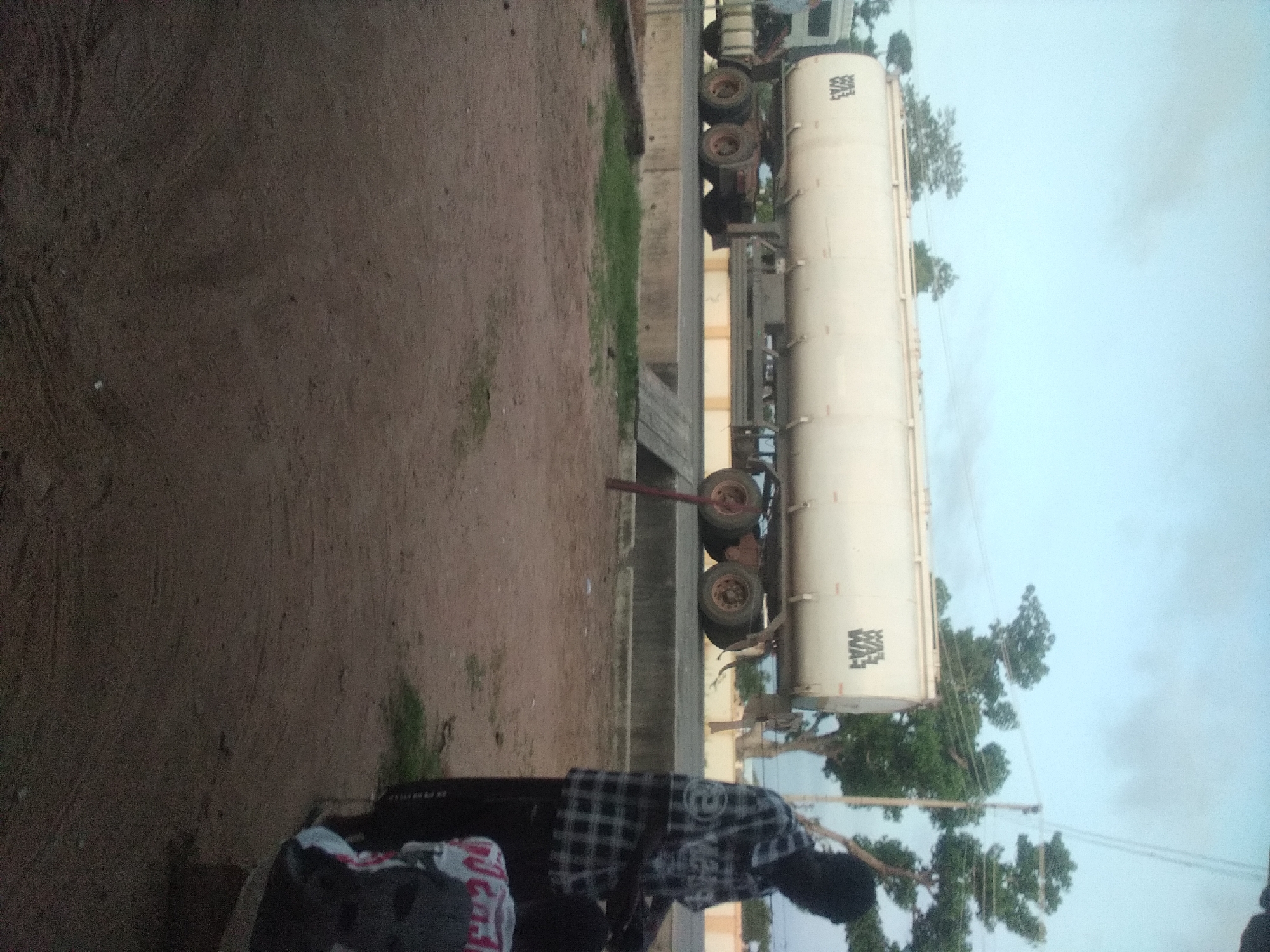 In Jalingo, Nigeria, a large white tanker truck is parked on a paved surface, possibly a road or a weighbridge, with two sets of wheels visible on its trailer. The truck has "WAL LAW" printed on its side in black lettering. To the right of the truck, a dark-skinned man wearing a black and white checkered shirt and dark pants stands facing away from the camera, holding a white object. Another person, partially visible, is in front of him, wearing a red and white patterned shirt. The ground in the foreground is dirt and gravel. The background shows trees and a pale, cloudy sky.