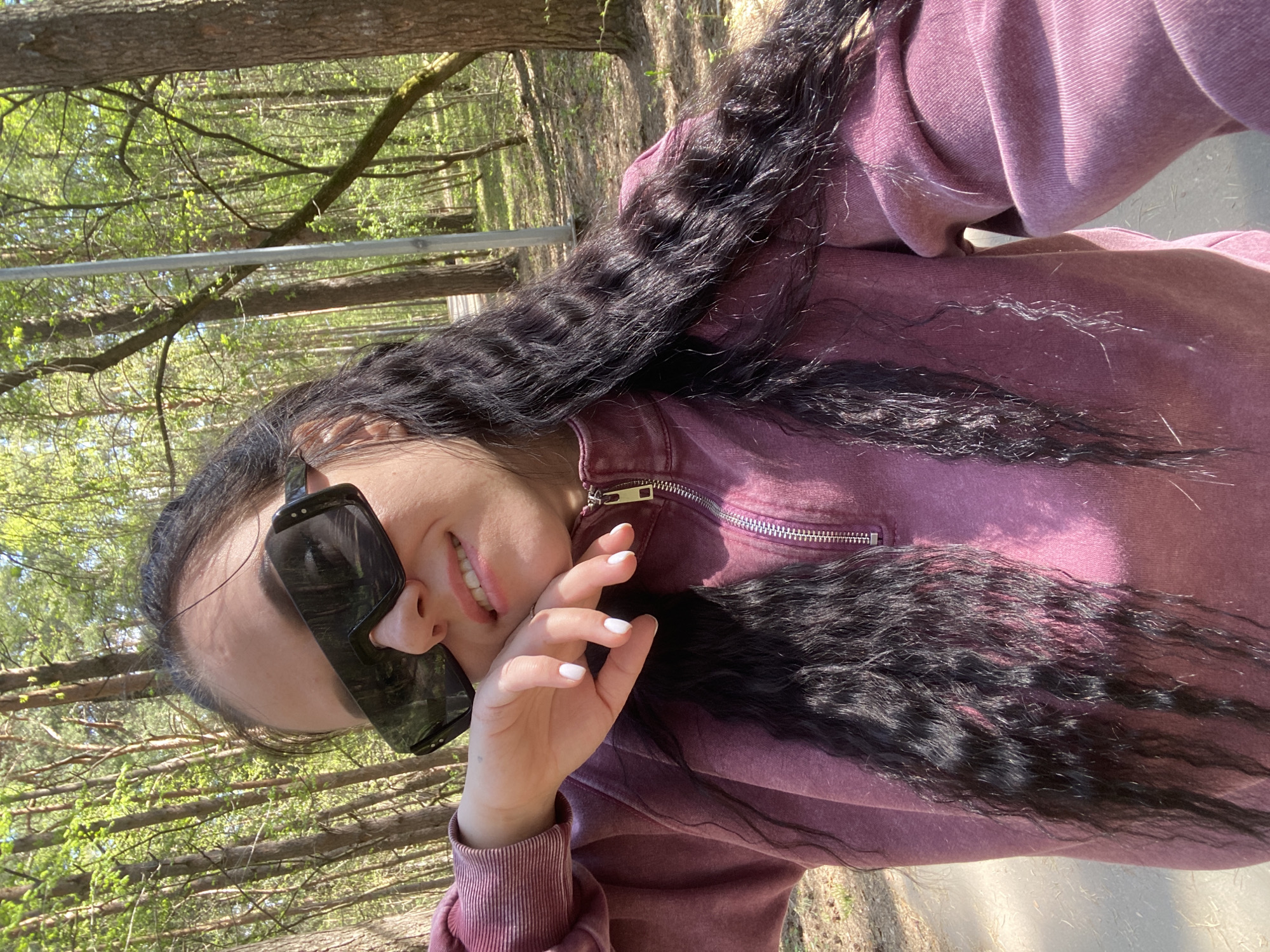 A young woman wearing sunglasses and a maroon sweatshirt poses for a selfie in a wooded area. The trees appear to be deciduous with new spring foliage. The location is potentially within the city of Perm, Russia, though no specific landmarks are visible to confirm this. The woman's hair is long, black, and wavy. She is smiling and holding her hand near her face. The background suggests a park or forest setting with trees and a path.