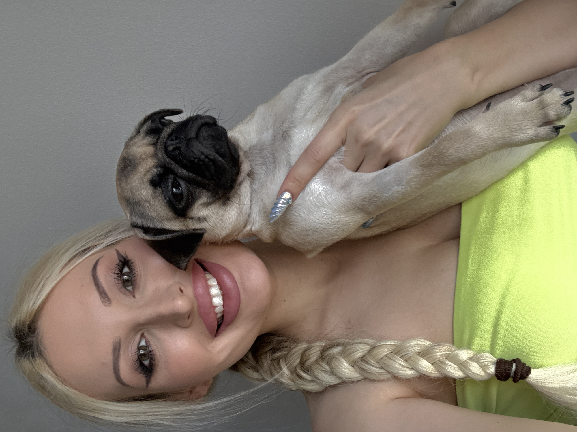A young blonde woman is holding a pug puppy in front of a plain gray wall. The woman has her hair in a braid and is wearing a bright green strapless top. She has on makeup, including winged eyeliner and pink lipstick, and her nails are decorated with a silver design. The pug puppy is light brown with a black face mask and is resting in the woman's arms. The image appears to be a selfie taken indoors, with no discernible features that would indicate a specific location in Podgorica, Montenegro.