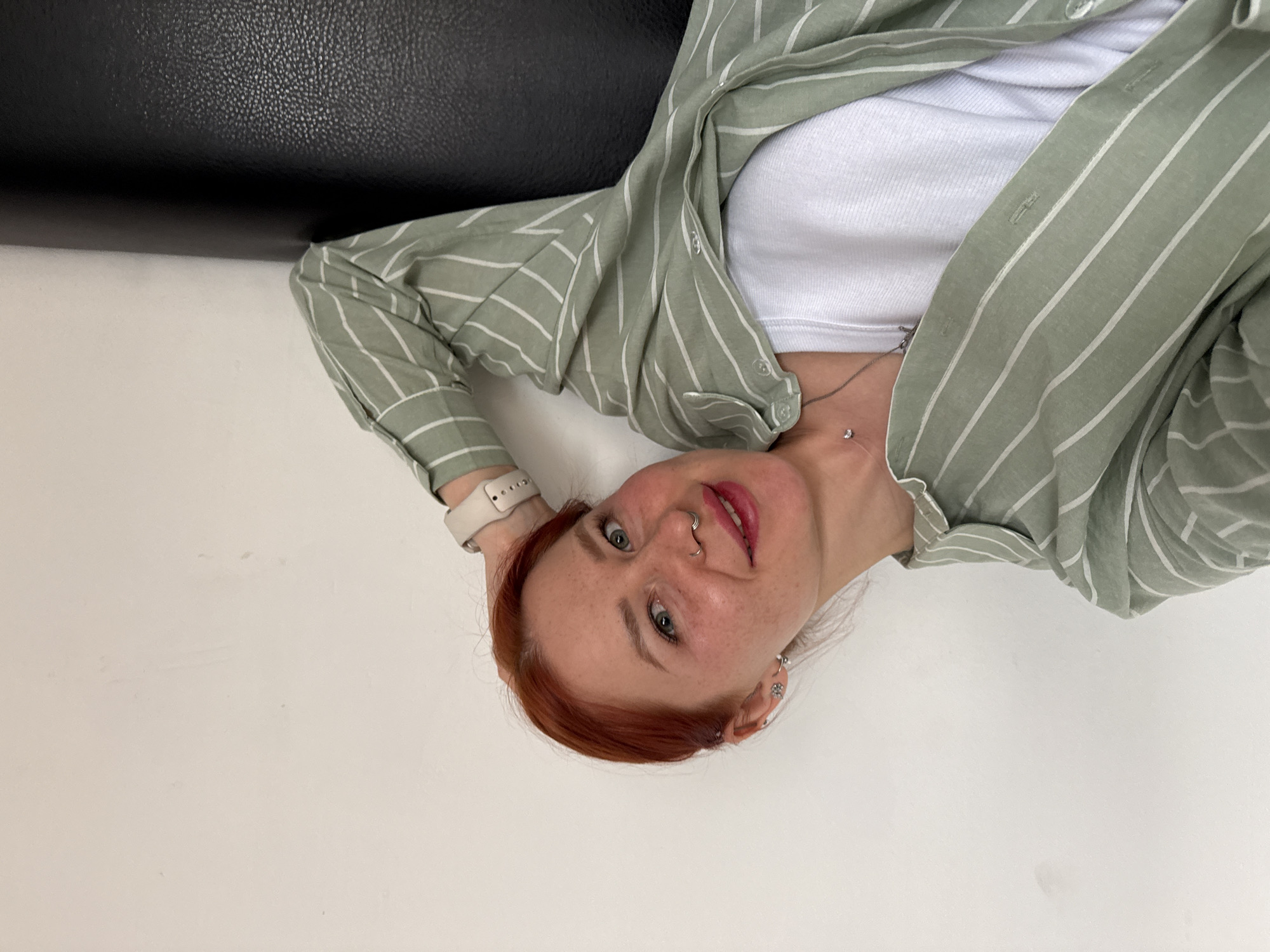 A young woman with reddish-brown hair and fair skin, adorned with freckles, is depicted in a close-up, horizontally oriented portrait. She has light-colored eyes (blue or green) and is smiling softly, looking directly at the viewer. Her left hand is placed behind her head, revealing a light-colored watch with a textured band on her wrist.
She is wearing a light green collared shirt with thin white vertical stripes, unbuttoned to show a white ribbed tank top underneath. A delicate silver necklace with a small, clear pendant rests on her chest. Facial piercings include a septum ring and a ring on her right nostril.
The setting appears to be indoors, with the woman positioned against two distinct surfaces. Behind her head and upper body is a dark, textured material, likely black leather or faux leather. The surface below her is plain and light-colored, suggesting she is reclining on a bed or sofa. The lighting is bright and even, illuminating her features. No visible text is present in the image.