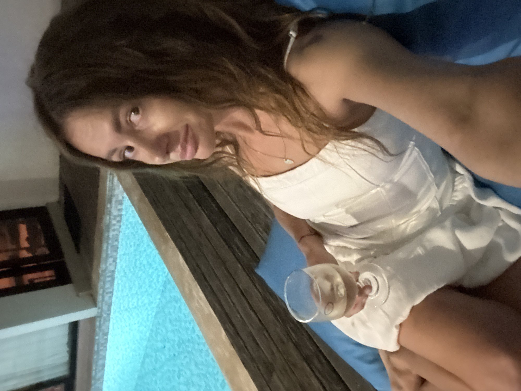 A woman with long brown hair and wearing a white dress is holding a glass of white wine. She is sitting in front of a window with a view of a pool. The location may be in the city of Kediri, Indonesia.