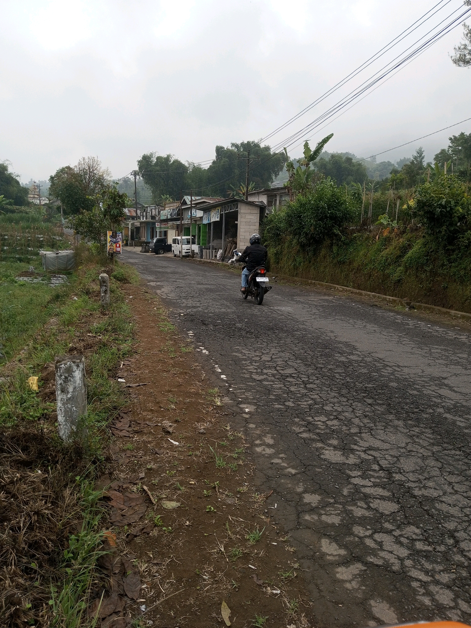 The image shows a road in Keseneng, Indonesia, on a cloudy day. A person is riding a motorcycle down the road. There are buildings on the side of the road, and trees and hills in the background. The road is cracked and appears to be in disrepair. There is vegetation on the left side of the road, and a few poles are visible. The sky is overcast, suggesting the possibility of rain or fog. The overall scene depicts a rural or semi-rural setting.