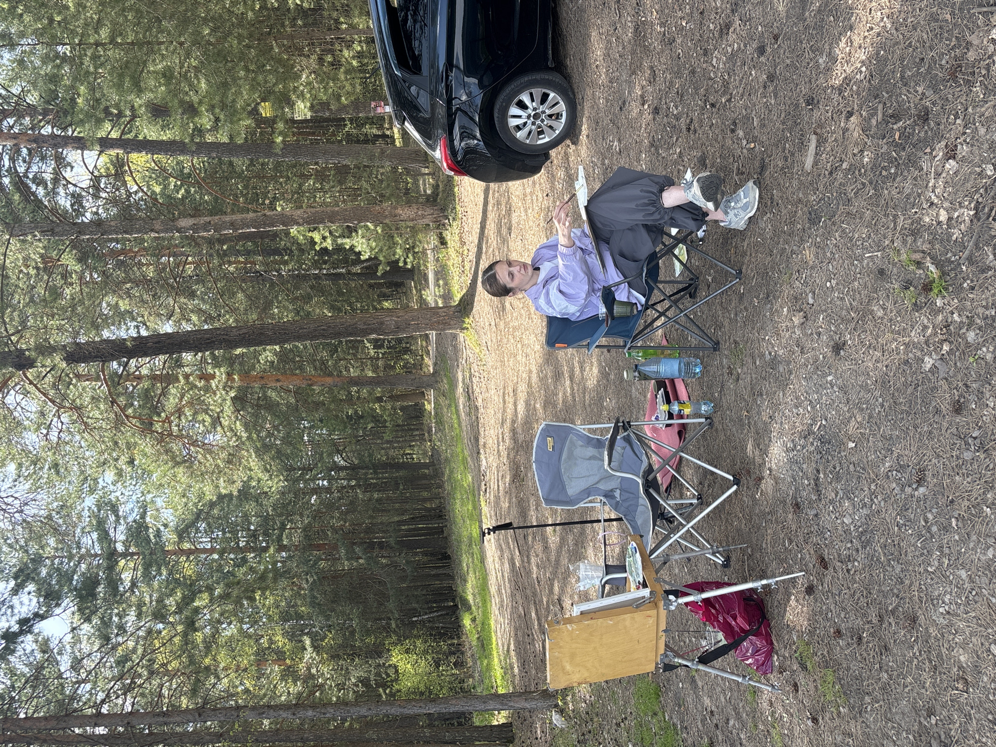 A fair-skinned woman sits in a blue folding chair in a sparse pine forest, a black car parked behind her. She wears a light purple jacket and grey pants, and appears to be painting on a canvas, which rests on a portable easel nearby. A second, empty grey folding chair is positioned next to the easel. The ground is covered in pine needles and cones. This location is likely within or on the outskirts of the city of Krasnoyarsk, Russia, due to the presence of pine forests.