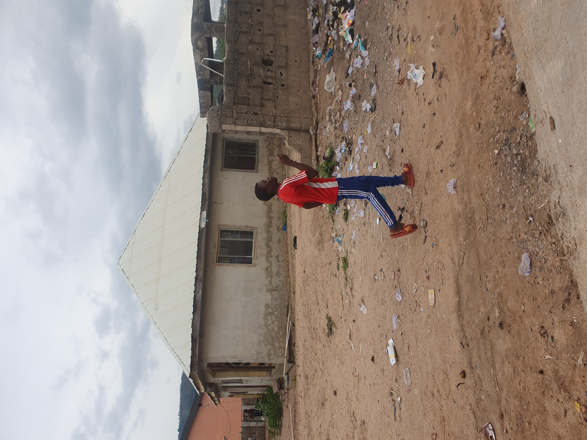 The image captures an outdoor scene in Lokoja, Nigeria, under an overcast sky filled with grey clouds. A young person, appearing to be male, is the central subject, standing on a dirt ground that is notably covered with scattered litter, including plastic bags, paper, and other debris. He is dressed in a red short-sleeved athletic shirt featuring white stripes on the sleeves, blue athletic pants with white stripes running down the sides, and red sandals or slides. He is looking upwards and to his right, with his right hand slightly raised as if gesturing or pointing towards something unseen.
In the background, to the left, stands a light-colored building with a white corrugated metal roof and two rectangular windows. Behind the person and further to the right, another structure, possibly incomplete or in disrepair, is visible, constructed from concrete blocks with some exposed elements. A portion of another building with a darker roof is also visible on the far left. The setting suggests a semi-urban or residential area with a mix of building types and noticeable ground pollution. The cloudy conditions indicate it is daytime. There is no visible text in the image.