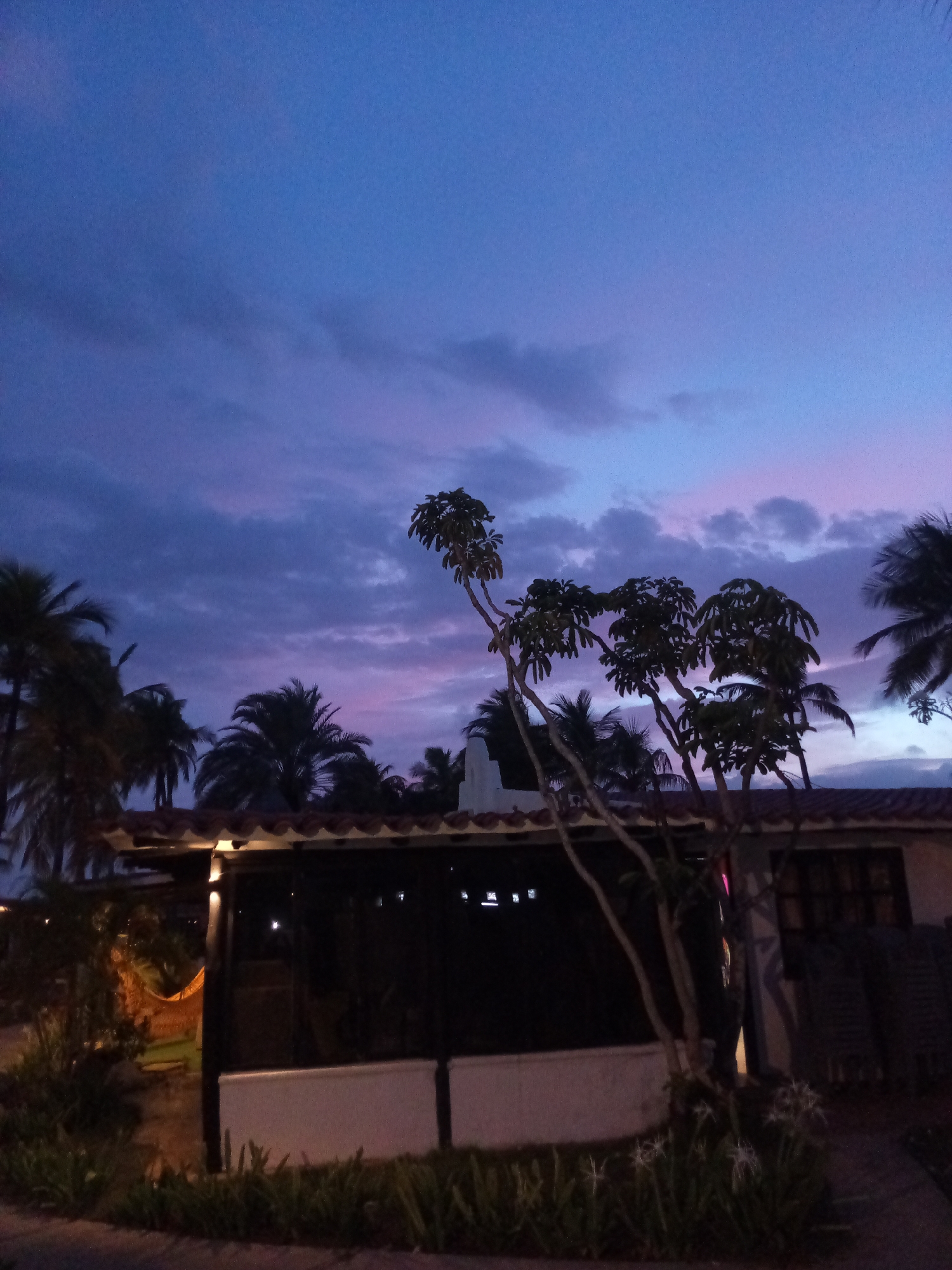 A photograph taken at dusk in Porlamar, Venezuela, captures a serene, twilight scene with a clear, gradient sky shifting from deep blue at the top to hues of pink and purple near the horizon. Scattered clouds drift across the sky, adding texture and depth to the view. In the foreground, a building with a tiled roof and open-air structure is partially visible, illuminated by soft, warm lights. Tropical palm trees, silhouetted against the colorful sky, surround the building, contributing to the exotic ambiance. A hammock is visible in the distance, suggesting a leisurely setting. The overall impression is one of tranquil beauty, characteristic of a tropical evening.