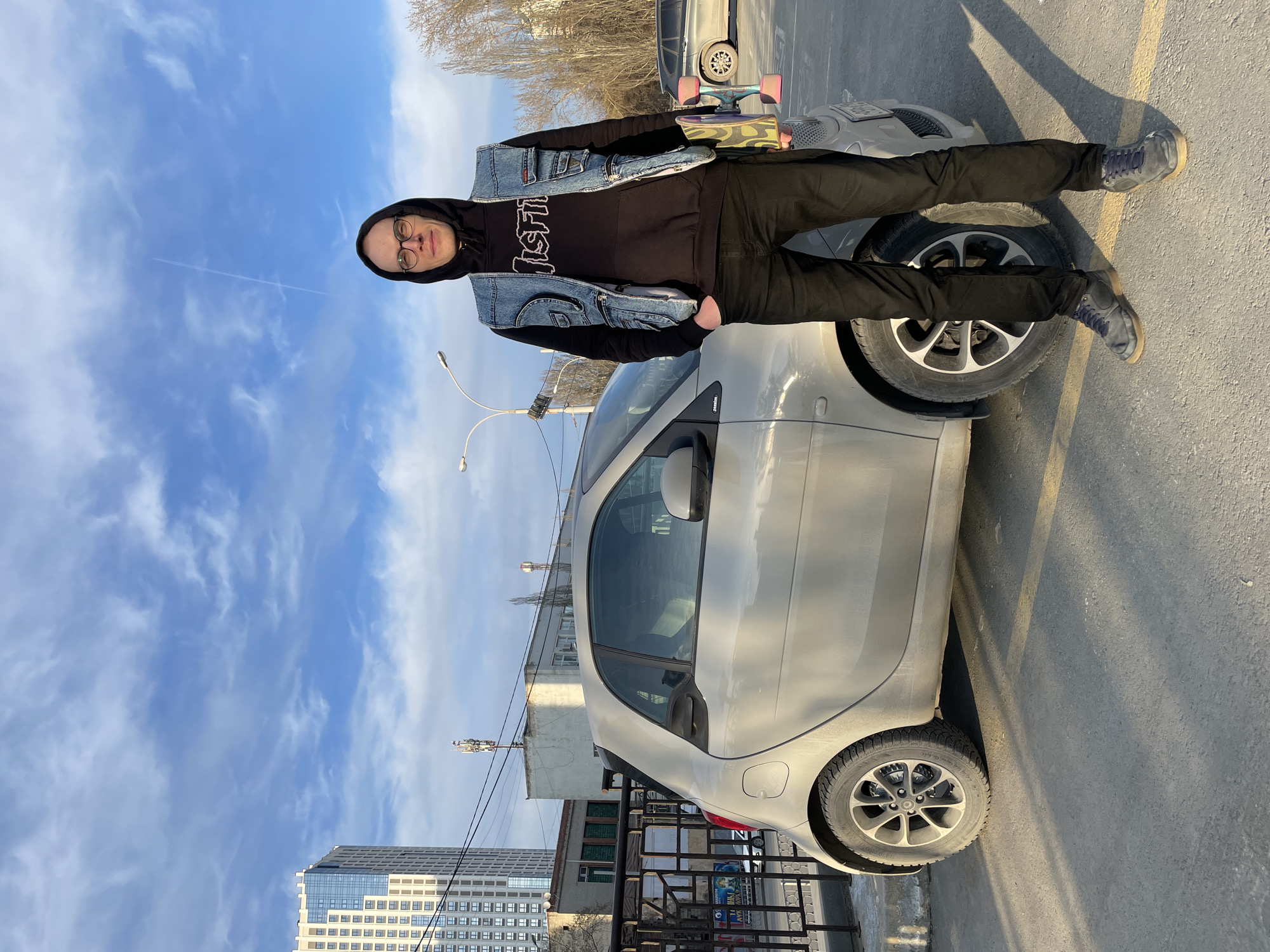 The image shows a man standing on the front wheel of a silver Smart car. He is wearing glasses, a black hooded sweatshirt with a graphic on the front, a denim vest, black pants, and blue sneakers. He is holding a skateboard in his left hand. The car is parked on an asphalt surface, possibly a street. Behind the car and the man, there is another car and a building, and in the background, there is a blue sky with scattered clouds. A tall modern building is visible in the distance. It's hard to determine the exact location in Yekaterinburg.