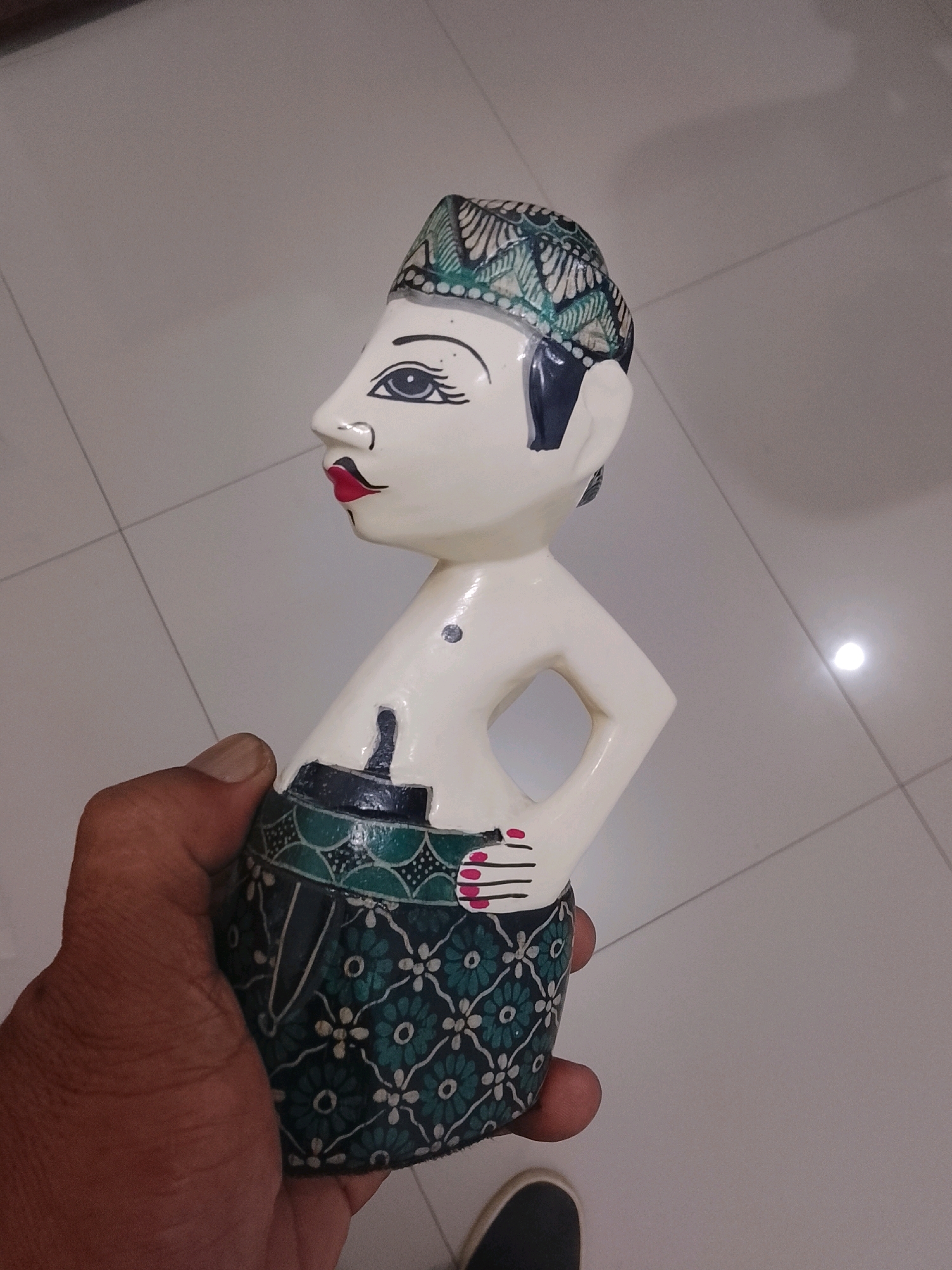 A hand holds a carved wooden puppet, likely a Wayang Golek, against a tiled floor in what appears to be an indoor setting in Slawi, Indonesia. The puppet is white with black and green painted details, featuring a traditional headdress with intricate patterns, a stylized face with red lips and defined eyes, and a skirt adorned with floral motifs. A dark blue and white patterned band encircles the waist of the puppet. The hand holding the puppet is brown-skinned. The floor is composed of large, light gray tiles with visible grout lines forming a grid pattern. A bright, circular reflection is present on the floor in the upper right portion of the image, indicating a light source.