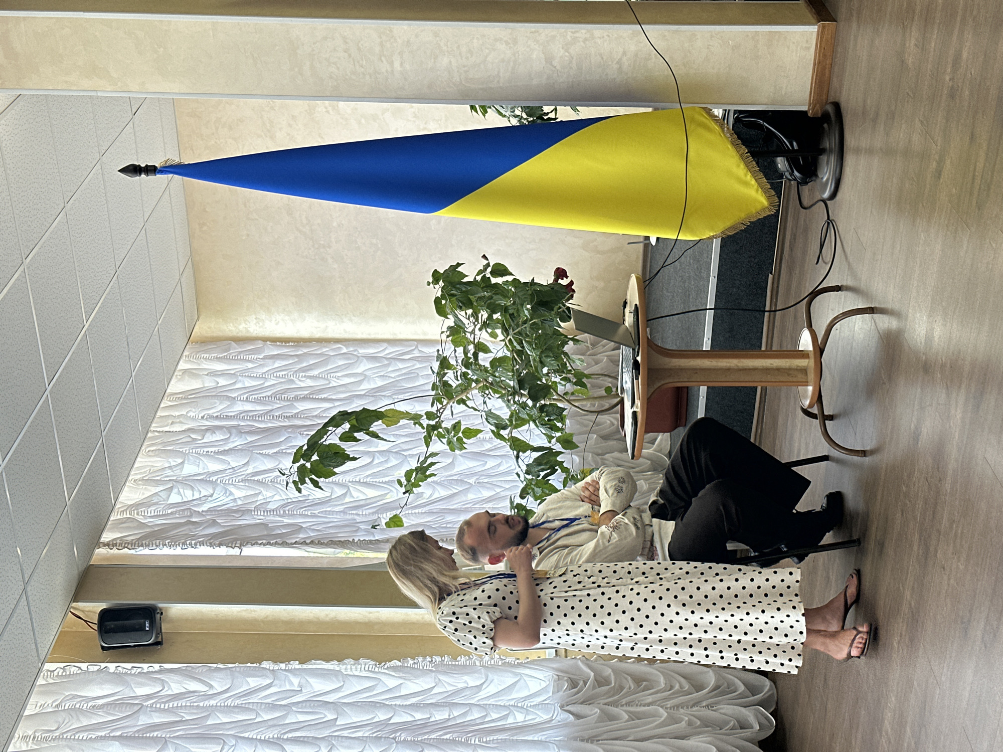 This media file captures an indoor scene featuring two individuals, a man and a woman, in what appears to be a formal or semi-formal setting, possibly a hall or meeting room, in Myronivka, Ukraine. The Ukrainian flag is prominently displayed, suggesting a patriotic or official context.
The man is seated, wearing a light-colored shirt and dark trousers. He has a beard and appears to be looking towards the woman. The woman, standing, is dressed in a white dress with black polka dots and is also facing the man, with her hands clasped. Their body language suggests an intimate or engaged conversation.
In the background, there is a plant with green leaves and a red flower, adding a touch of nature to the indoor environment. The room is decorated with white drapes that have a textured pattern. A speaker is mounted on the wall. The lighting suggests it is daytime. The overall mood appears to be calm and conversational. No visible text is present in the image.