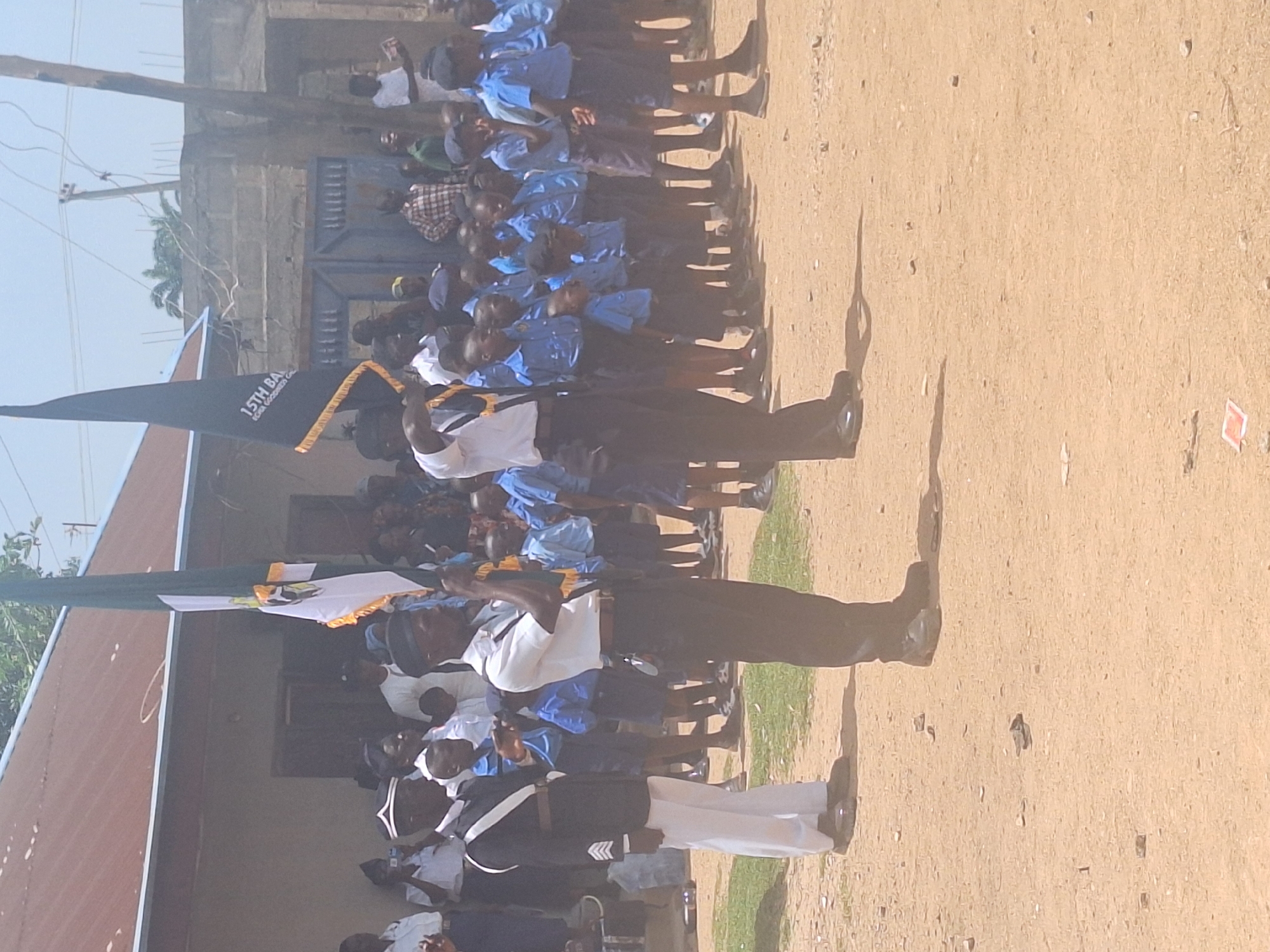 A group of people is gathered outdoors on a sandy surface in Sabon Tasha, Nigeria. In the foreground, three men in uniforms stand in a row; one in a black and white uniform and two in white shirts and black pants. These men are holding flags. Behind them is a line of children wearing blue shirts, and several adults can be seen standing amongst them. A building is visible in the background.