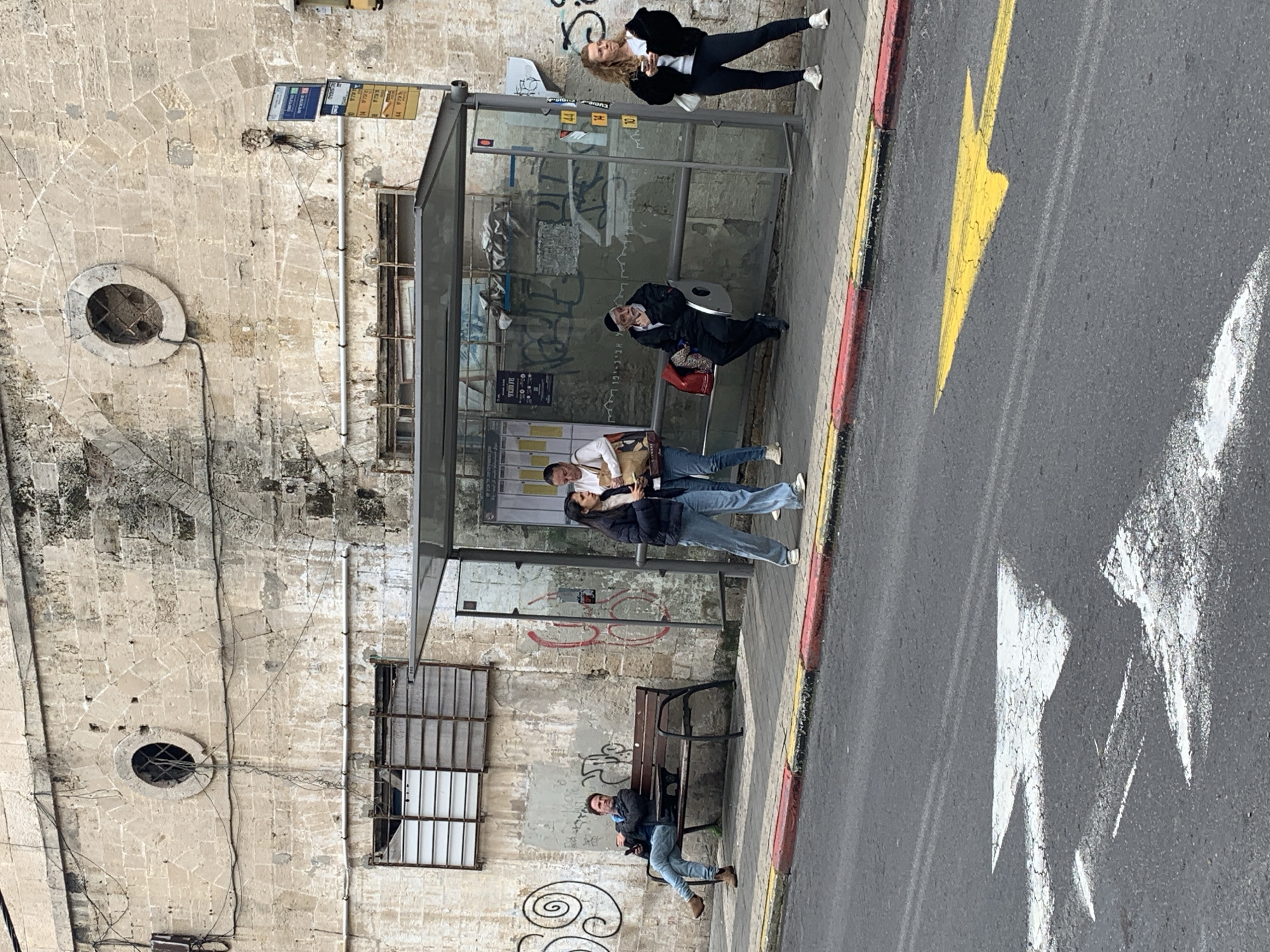 The image depicts a street scene in Tel Aviv, Israel. Several people are visible; one stands near a bus shelter, while another sits inside it. Two more individuals stand near the bus shelter, and another sits on a bench. The environment includes a wall with windows, round openings, and exposed wiring. A yellow arrow is painted on the street.