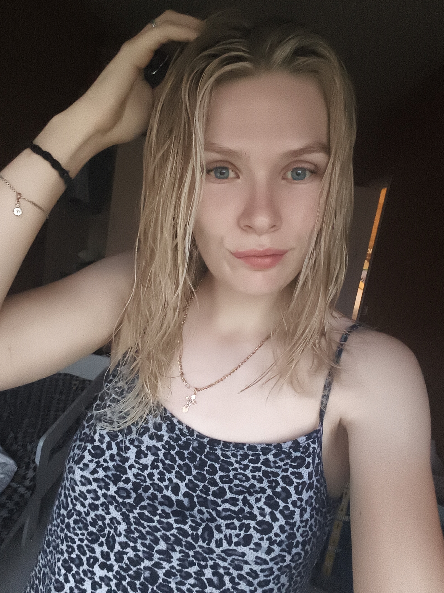 A young woman with blonde hair and blue eyes, wearing a leopard print top and necklaces, is captured in a close-up selfie. Her left arm is raised, with her hand touching her hair, and she is wearing bracelets on her left wrist. The background is dark and indistinct, suggesting an indoor setting with minimal lighting. The image appears to be taken in the city of Русский Вожой, Russia.