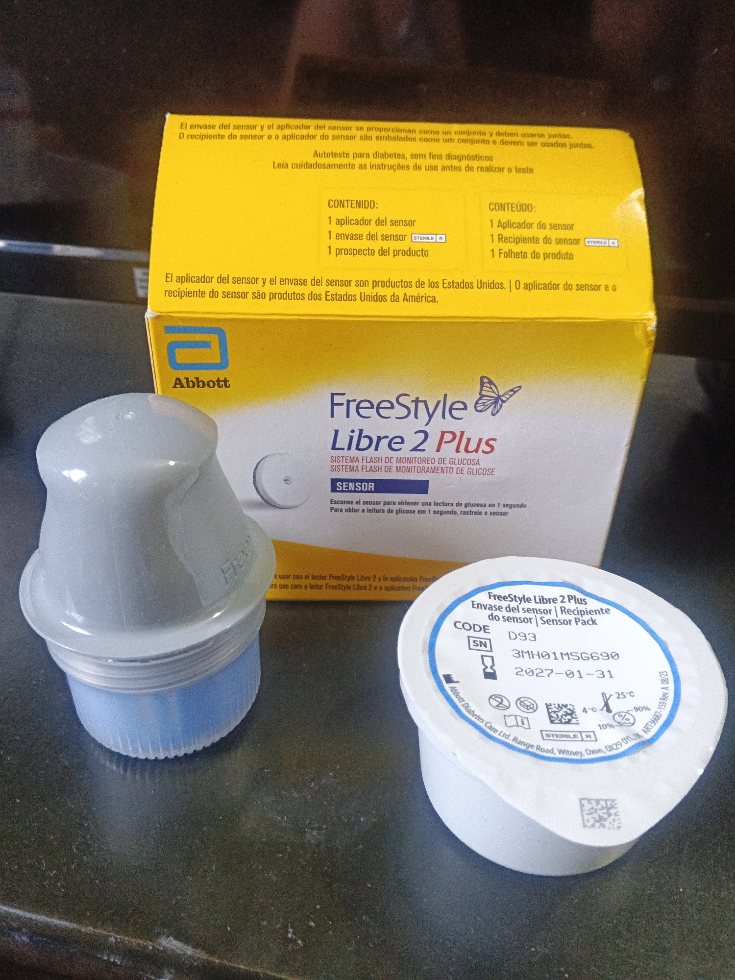 The image displays a FreeStyle Libre 2 Plus glucose monitoring system. A yellow box with "FreeStyle Libre 2 Plus" printed on it is in the background. In the foreground are two white and blue containers: a sensor applicator on the left, and a sensor pack on the right. The sensor pack has information printed on it including the code D93, serial number 3MH01M56690, and an expiration date of 2027-01-31. The packaging is labeled in both Spanish and Portuguese. The image was probably taken indoors on a dark surface. There is no information to determine the location of the image in Caracas, Venezuela.