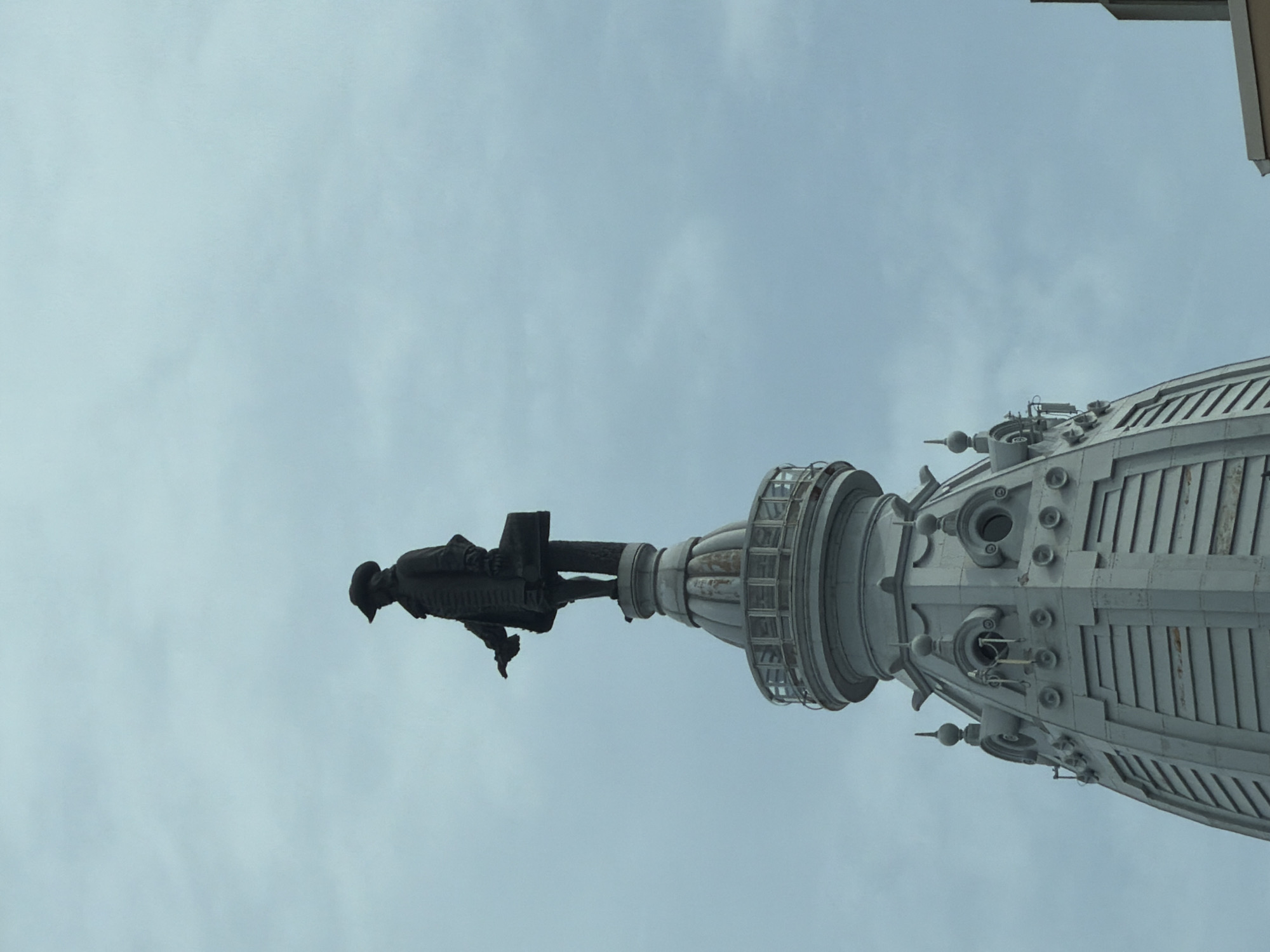 The image shows a low-angle, exterior shot of the statue of William Penn atop Philadelphia City Hall in Philadelphia, Pennsylvania, United States of America. The statue, made of bronze, is silhouetted against a cloudy, grey sky. Penn is depicted wearing a tricorn hat and a long coat, holding a scroll in his left hand and a sword in his right. He stands on a decorative pedestal which is part of the building's dome. The dome itself is ornate, with multiple levels and architectural details, including what appear to be windows or decorative elements. The statue and dome are predominantly grey. The overall impression is one of historical grandeur and a sense of looking up at a significant landmark.