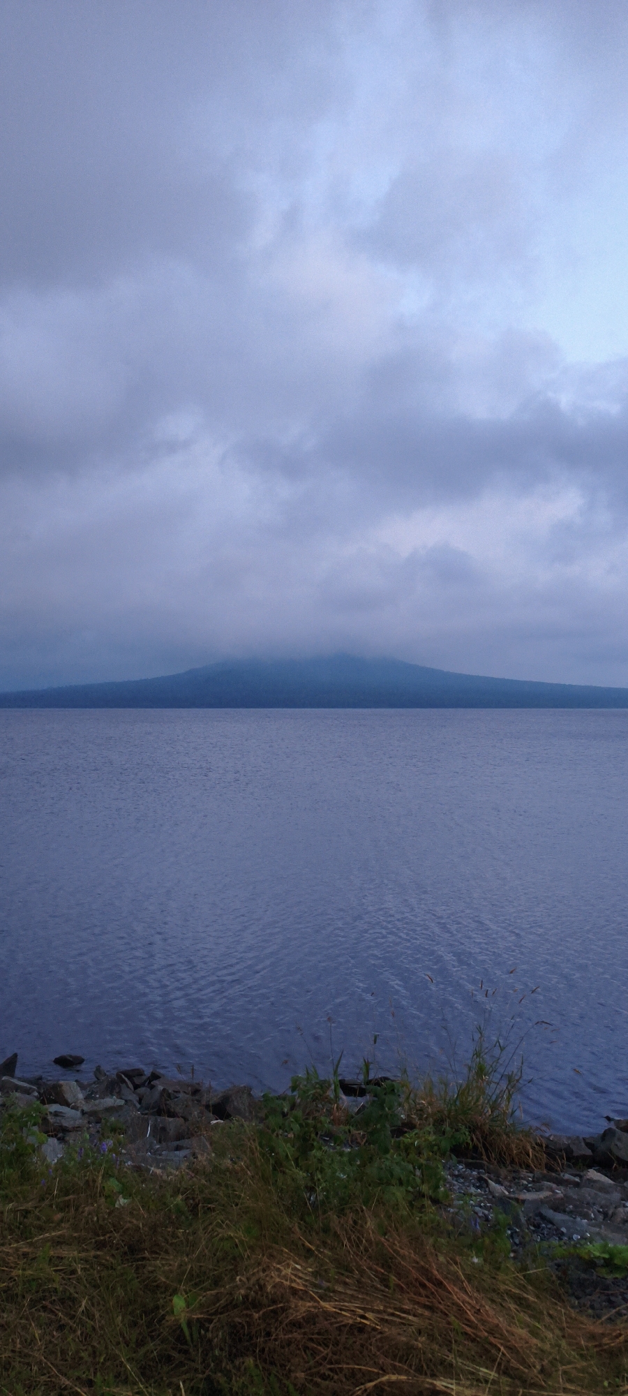 The image shows a calm, wide body of water, likely a lake, under an overcast sky. In the distance, a large, rounded mountain is visible, partially shrouded in mist near its peak. The foreground consists of a rocky shoreline with dry grasses and some green foliage. The overall atmosphere is subdued and serene, with muted colors dominated by blues, grays, and browns. The location is specified as the city of Zyuratkul', Russia.