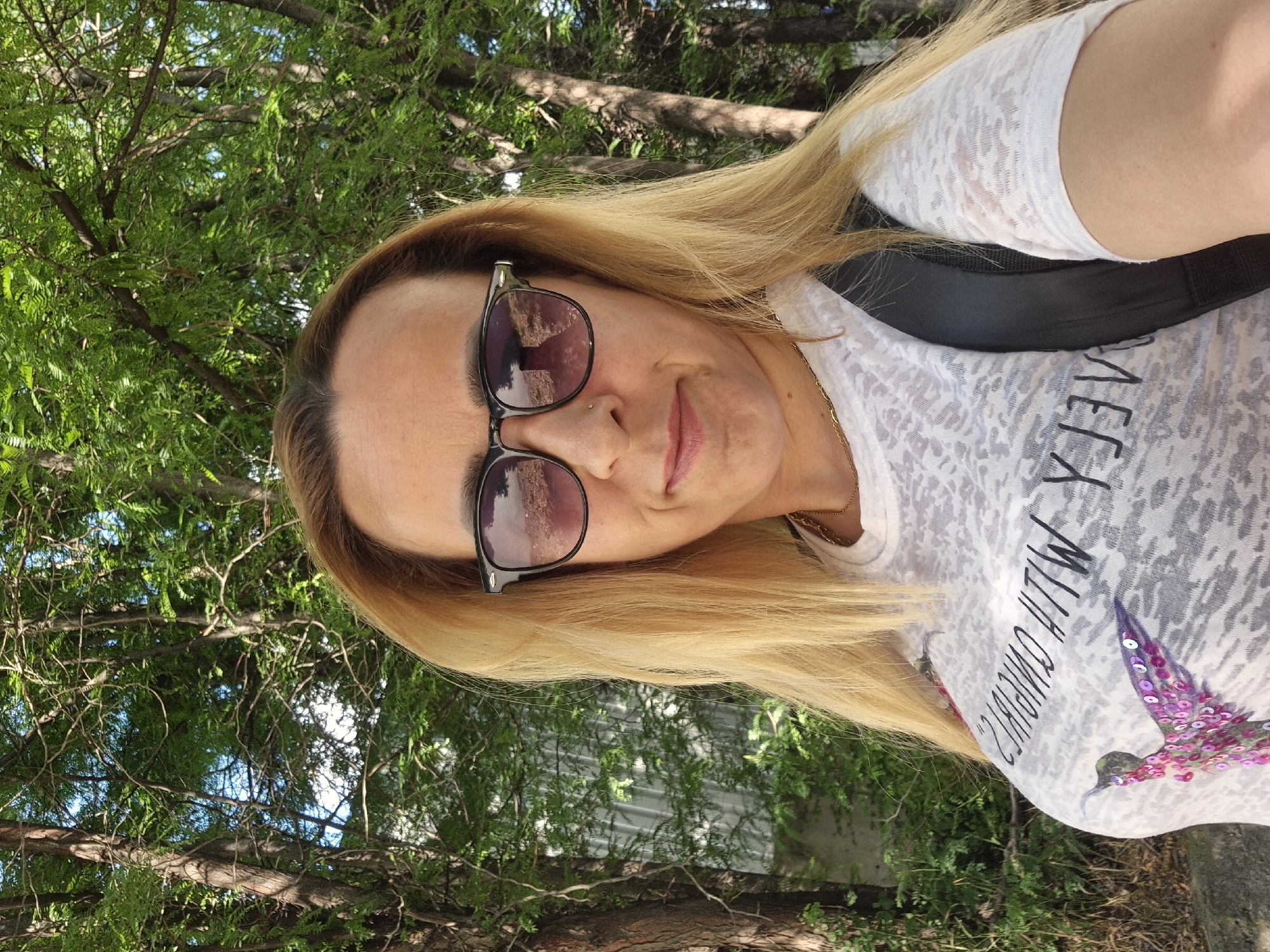 In a medium shot taken outdoors, a blonde woman with sunglasses and a white t-shirt is looking at the camera. The t-shirt has a colorful hummingbird graphic and text that reads "MY CRUSHES WITH?". She has blonde hair that is parted to the side and reaches her shoulders. She is wearing dark sunglasses with purple lenses, which reflect the surroundings. The background consists of trees with green foliage and some branches. The woman is wearing a thin gold chain necklace. The image appears to be a selfie taken outdoors, possibly in a park or a wooded area.