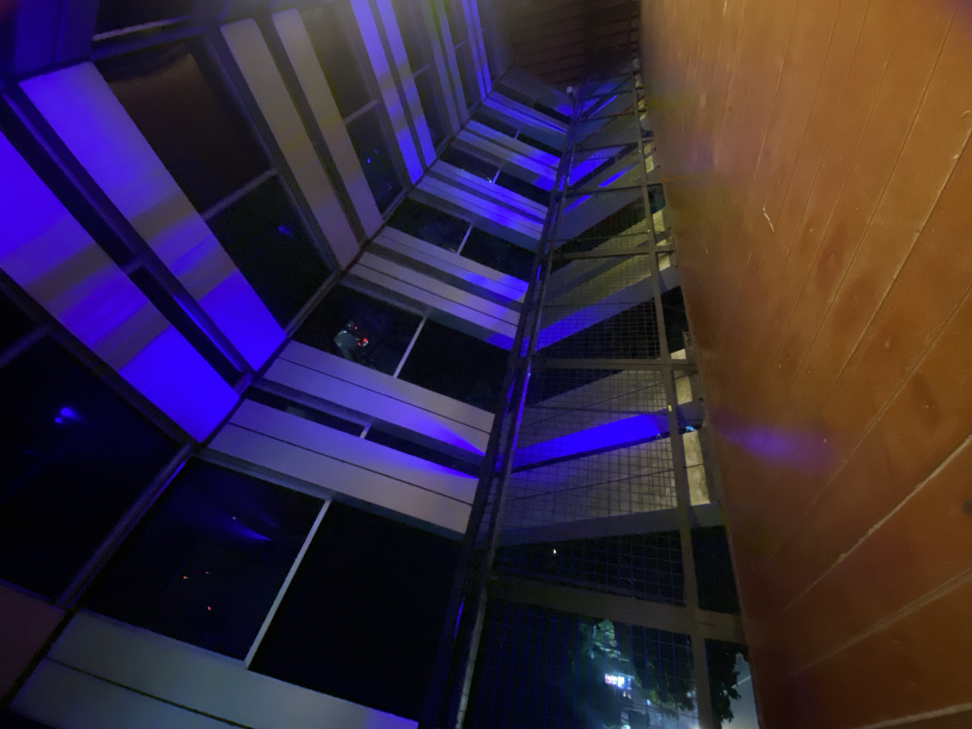 The media file captures an upward view of a modern building's exterior at night. The building features a facade with alternating panels of light-colored material and dark windows. Blue LED lights are illuminating some of the panels, creating a striking visual effect. Adjacent to this facade is a metal grate structure, possibly a walkway or part of the building's internal scaffolding, also lit by the blue lights in sections. To the right, a portion of a wooden-paneled wall is visible, its texture and color suggesting an outdoor setting or a transition to another architectural element.
The scene is set at night, as indicated by the darkness and the artificial lighting. There are no clear subjects like people or distinct objects, but reflections in the dark windows hint at the surrounding environment or interior lights. The overall mood is contemporary and somewhat dramatic due to the intense blue lighting against the dark backdrop. Location cues like "Banjarbaru, Indonesia" are provided but are not visually discernible within the image itself. No visible text is present in the media file.
