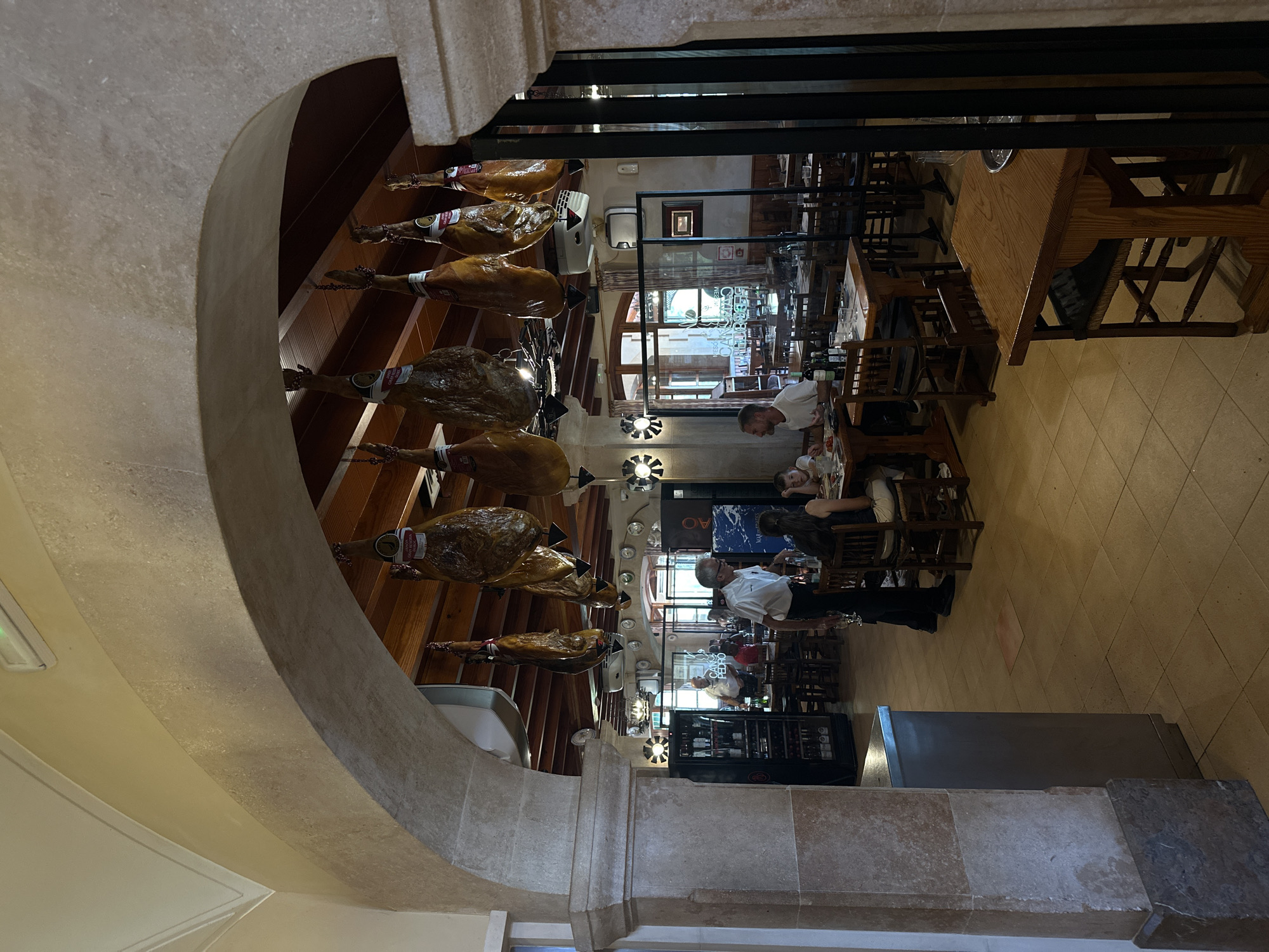 The image depicts the interior of a restaurant, likely located in Palma de Mallorca, Spain, a common misconception being Gènova which is a different region. The scene is framed by a stone archway, with several legs of cured ham hanging from the ceiling. Inside, diners are seated at wooden tables, some interacting with each other and others with staff. A waiter in a white shirt is seen serving at one table, while other patrons, including men, women, and children, are engaged in conversation or eating. The restaurant features a rustic aesthetic with wooden beams on the ceiling, tiled floors, and traditional wooden furniture. A glass-enclosed wine display is visible on the left side of the image, and ambient lighting creates a warm atmosphere. The overall impression is that of a traditional Spanish dining establishment.
