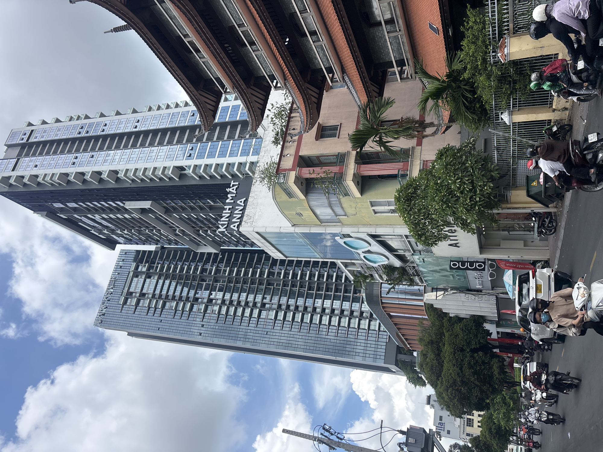 A street scene in a city, possibly Southeast Asia, features a mix of modern high-rise buildings and older, traditional architecture. Two very tall, modern glass skyscrapers dominate the background, their facades reflecting the cloudy blue sky. To the right, a multi-tiered traditional building with a dark-tiled, curved roof and ornate detailing stands in front of the skyscrapers. In the mid-ground, a narrower, colorful building with balconies and a palm tree is visible. The street level is busy with motorbikes, some carrying multiple passengers, and a few cars. Pedestrians are seen on the sidewalks, and storefronts are present, including one with a large glasses logo. The overall impression is a bustling urban environment where contemporary and traditional elements coexist.