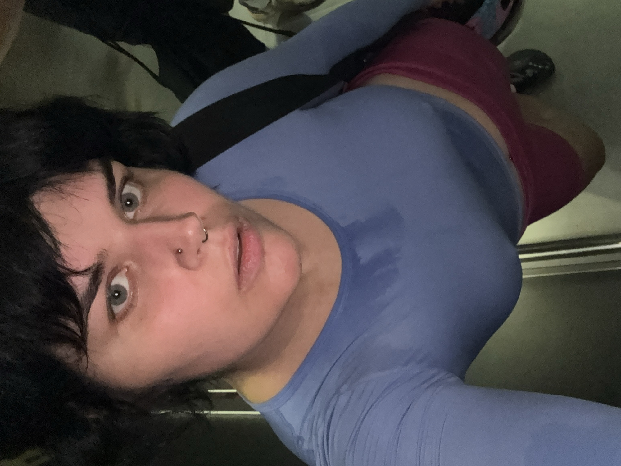 A close-up, high-angle shot shows a person wearing a blue long-sleeved shirt and pink shorts. The shirt appears damp in the chest area. The person has dark hair, light-colored eyes, and a nose ring. They are looking upwards, with their mouth slightly open. A backpack strap is visible over their shoulder. The background is dark and indistinct, with some vertical lines suggesting a doorway or elevator interior. The location is identified as Nong Pa Khrang, Thailand, but no visual elements in the image confirm this specific location.