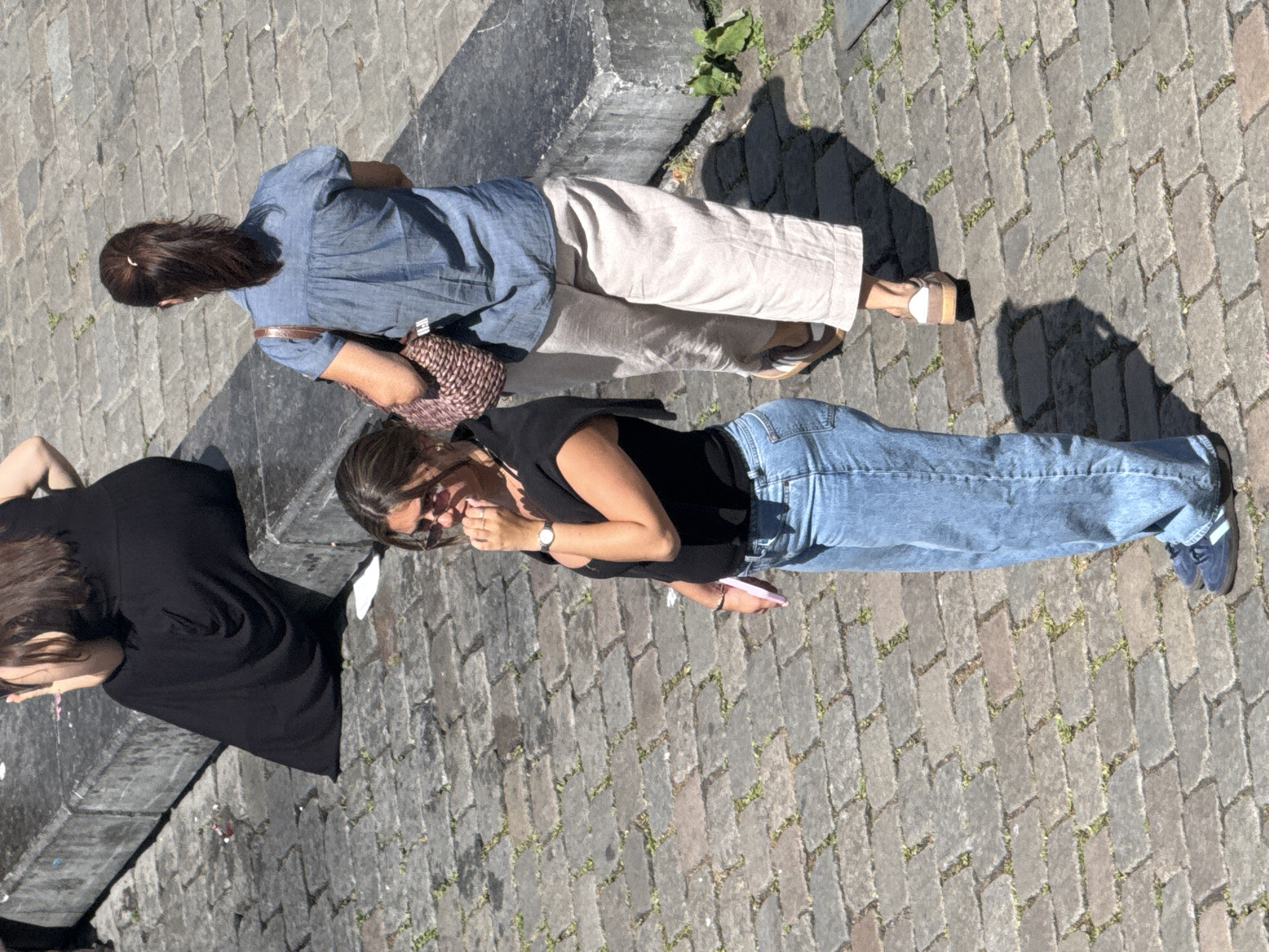 In Ghent, Belgium, a high-angle shot captures three women on a cobblestone street. In the foreground, a woman in a black shirt and blue jeans stands with her hand to her mouth, looking downwards. To her right, a woman in a blue denim shirt and tan trousers walks away from the camera, carrying a woven bag over her shoulder. In the background, a third woman in a long black dress sits on a concrete ledge, her back to the camera. The scene is lit by bright sunlight, casting distinct shadows on the cobblestones.