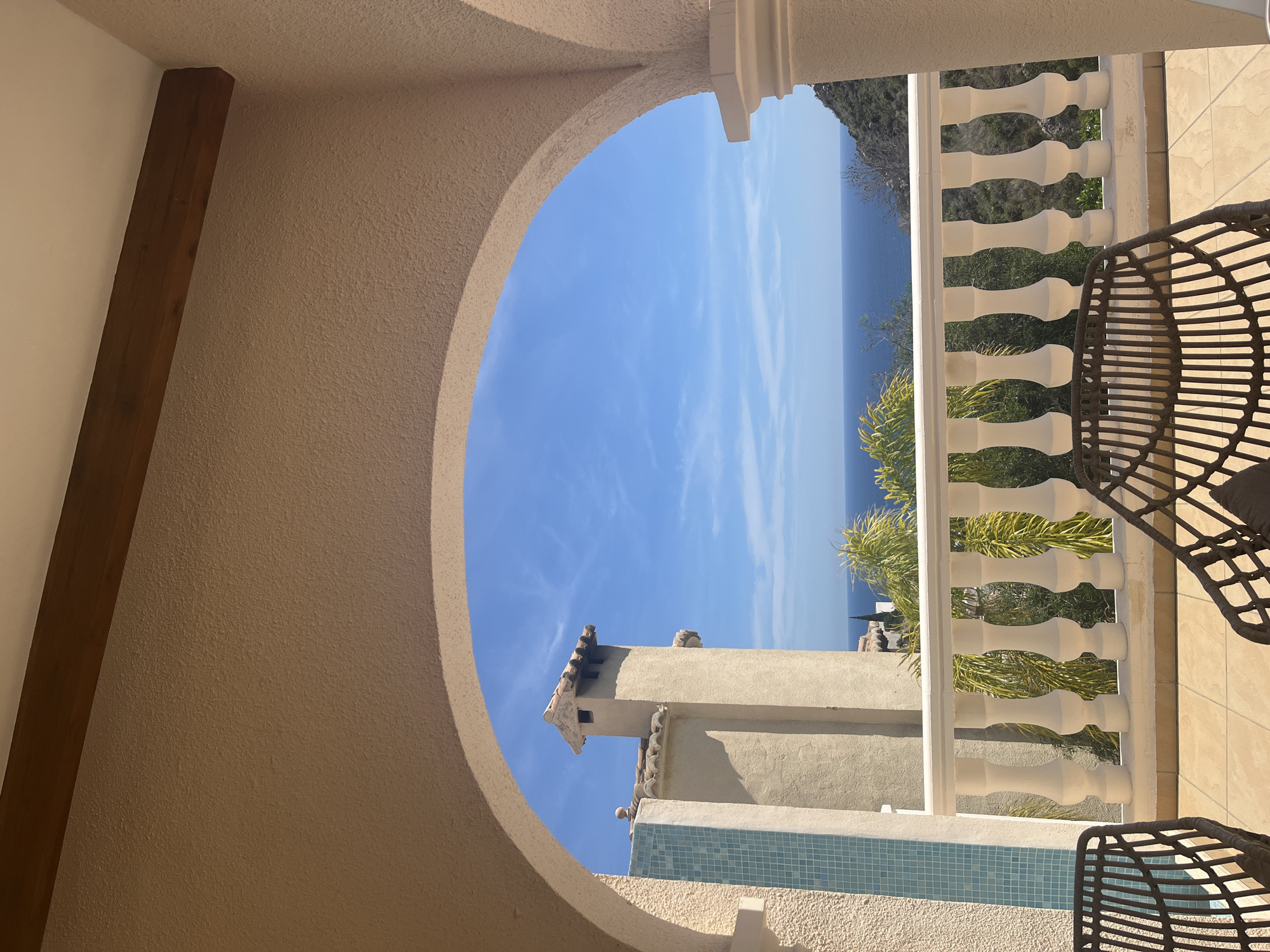 The image shows a view from an arched opening, possibly a window or doorway, overlooking a coastal scene in El Poble Nou de Benitatxell / Benitachell, Spain. The frame of the opening is made of a beige-colored stucco. A dark wooden beam is visible on the left side of the frame. Outside, there is a white balustrade or railing, partially obscuring a view of the Mediterranean Sea and the adjacent coastline. Vegetation, likely coastal plants and trees, is visible between the railing. A chimney is also partially visible below the horizon of the Mediterranean. A wicker chair is partially within the frame. The sky is clear and blue with faint clouds.