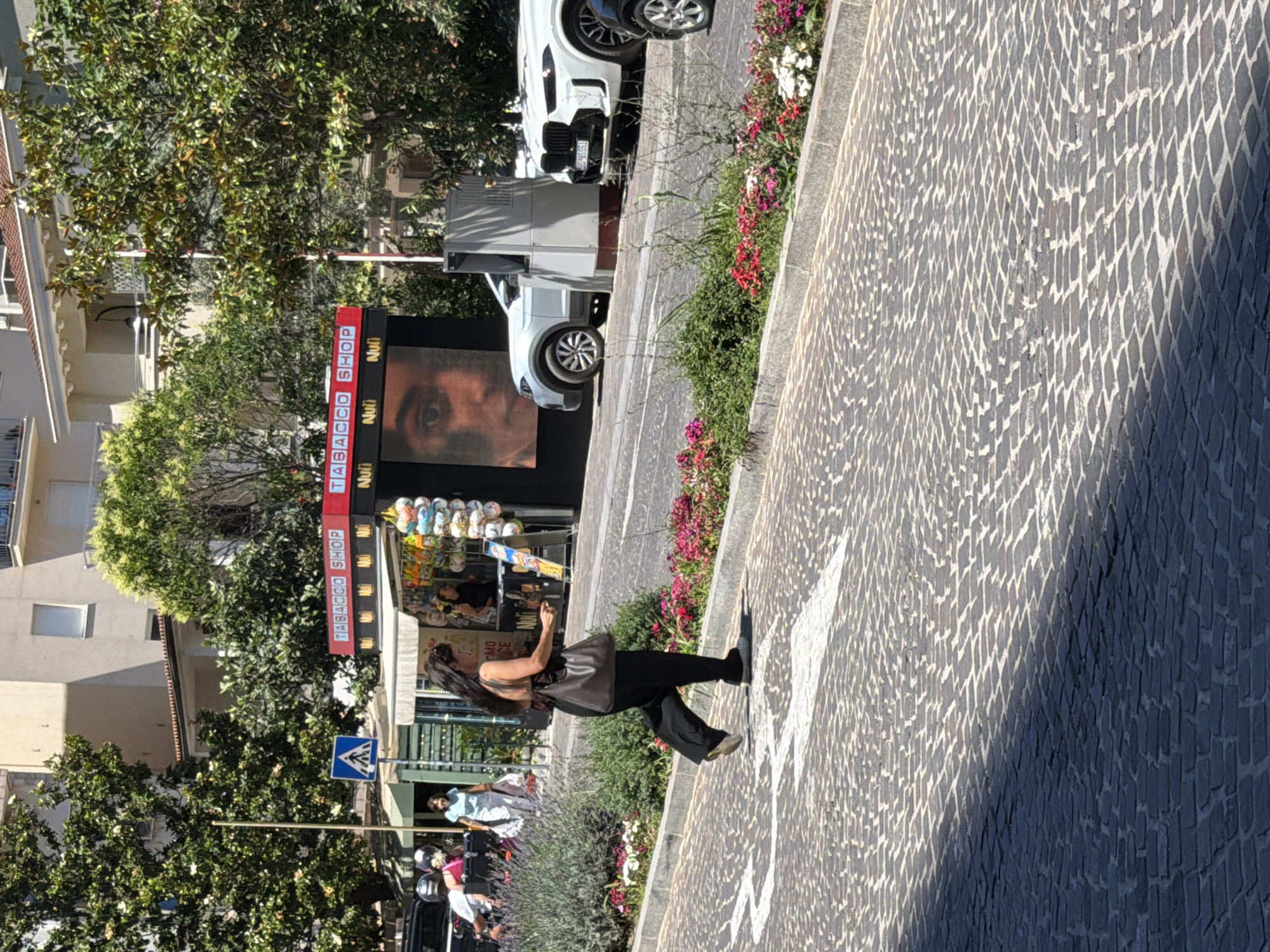 In the city of Budva, Montenegro, on a sunny day, a woman with long dark hair is walking on a cobblestone street. She is wearing a dark sleeveless top, black pants, and carrying a dark brown shoulder bag. She is walking towards the right side of the frame, away from a tobacco shop with a red awning and a large screen displaying a close-up of a person's eye. Behind the tobacco shop, a white SUV is parked. In the foreground, a patch of flowers and greenery lines the street. A white arrow is painted on the cobblestones, indicating a direction of travel. In the background, buildings and trees are visible.