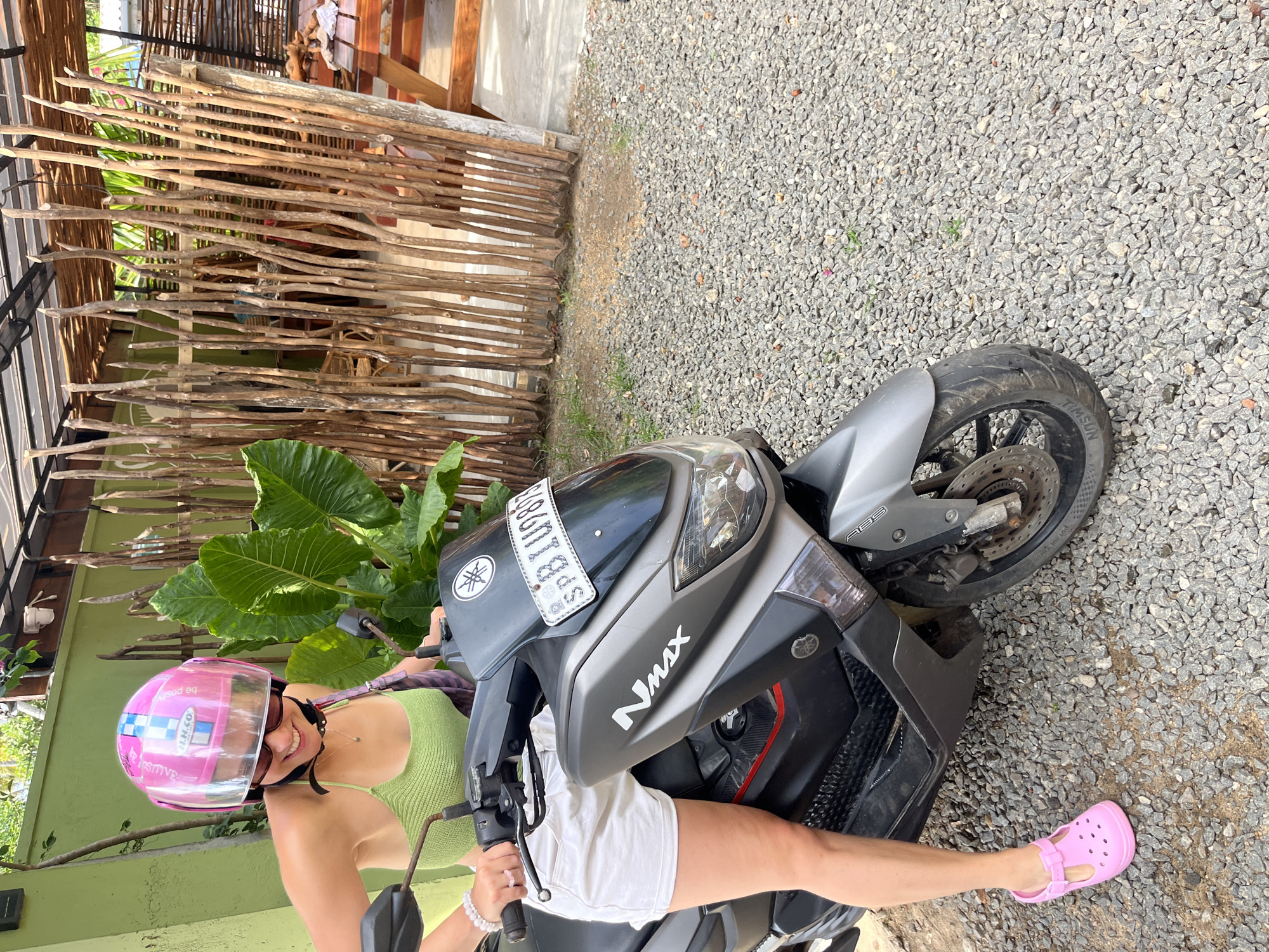 A woman wearing a pink helmet and a green top is sitting on a grey scooter in Kabalana, Sri Lanka. She is smiling and looking towards the camera. The scooter has a license plate with the letters "SPB". The background features a wooden fence and lush green foliage. The ground is covered in small grey pebbles.